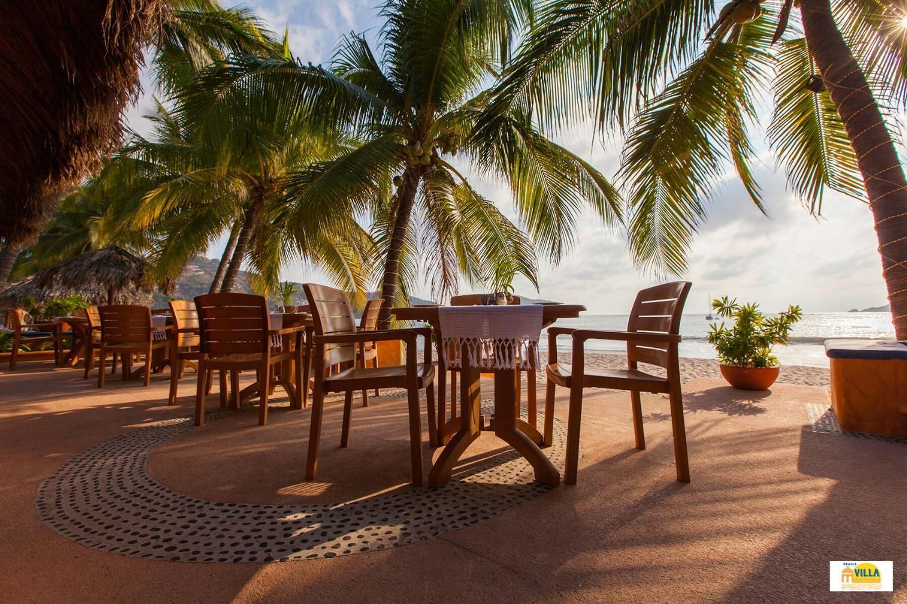 Una veranda fronte spiaggia con sedie e tavolo di legno, palme e vista sull'oceano.