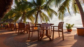 A beachside patio with wooden chairs and a table, palm trees, and a view of the ocean.