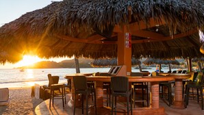 Un área de comedor al aire libre con sombrillas de paja, sillas de mimbre y vistas al océano al atardecer.