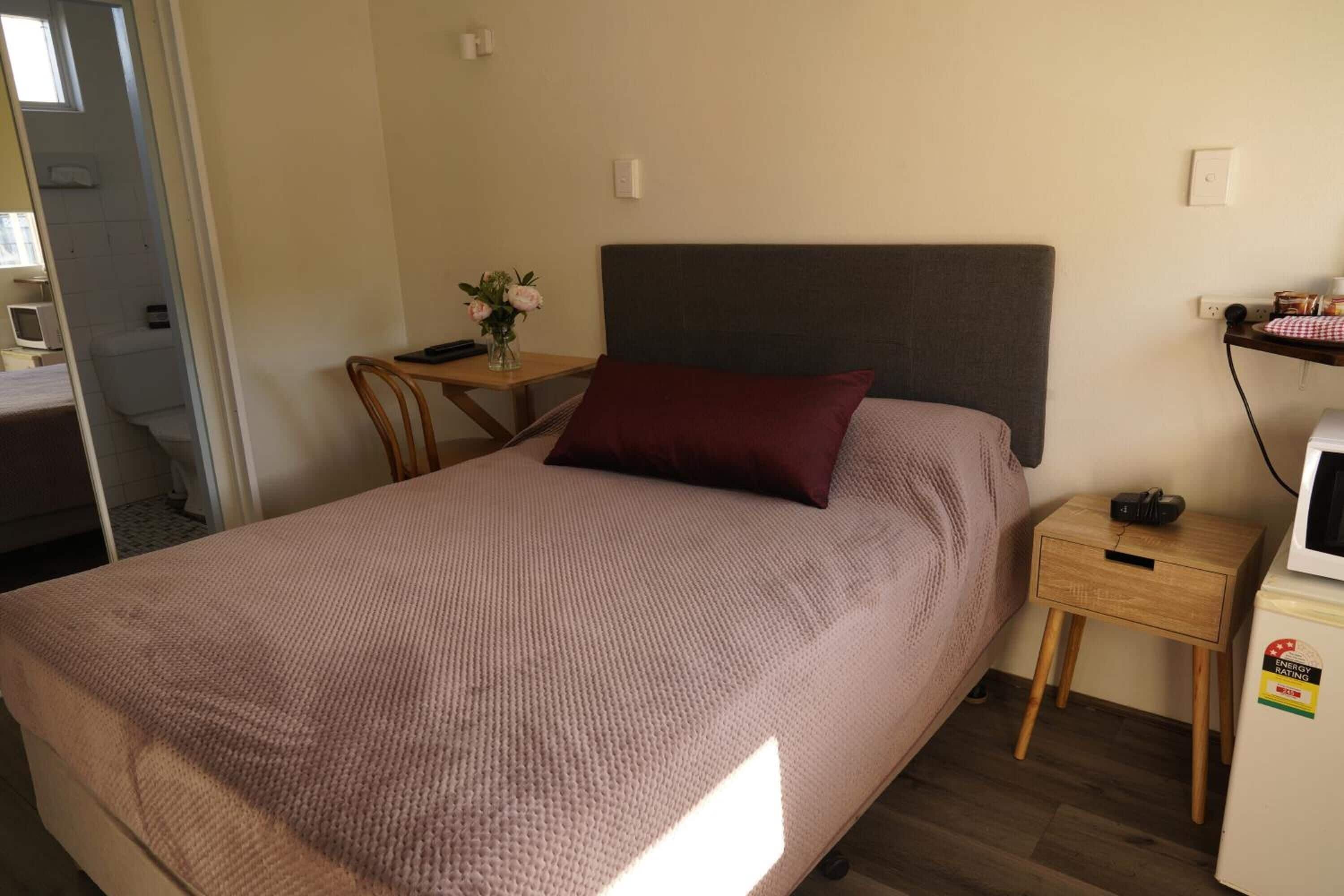 Une chambre à coucher comprenant un lit, une table de chevet, une petite table avec une chaise, une étagère fixée au mur et un réfrigérateur.