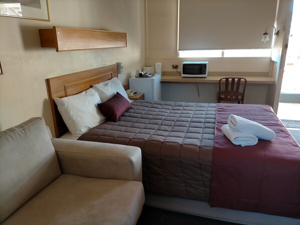 Quarto de hotel com cama, sofá, escrivaninha e computador, além de janela com persianas.