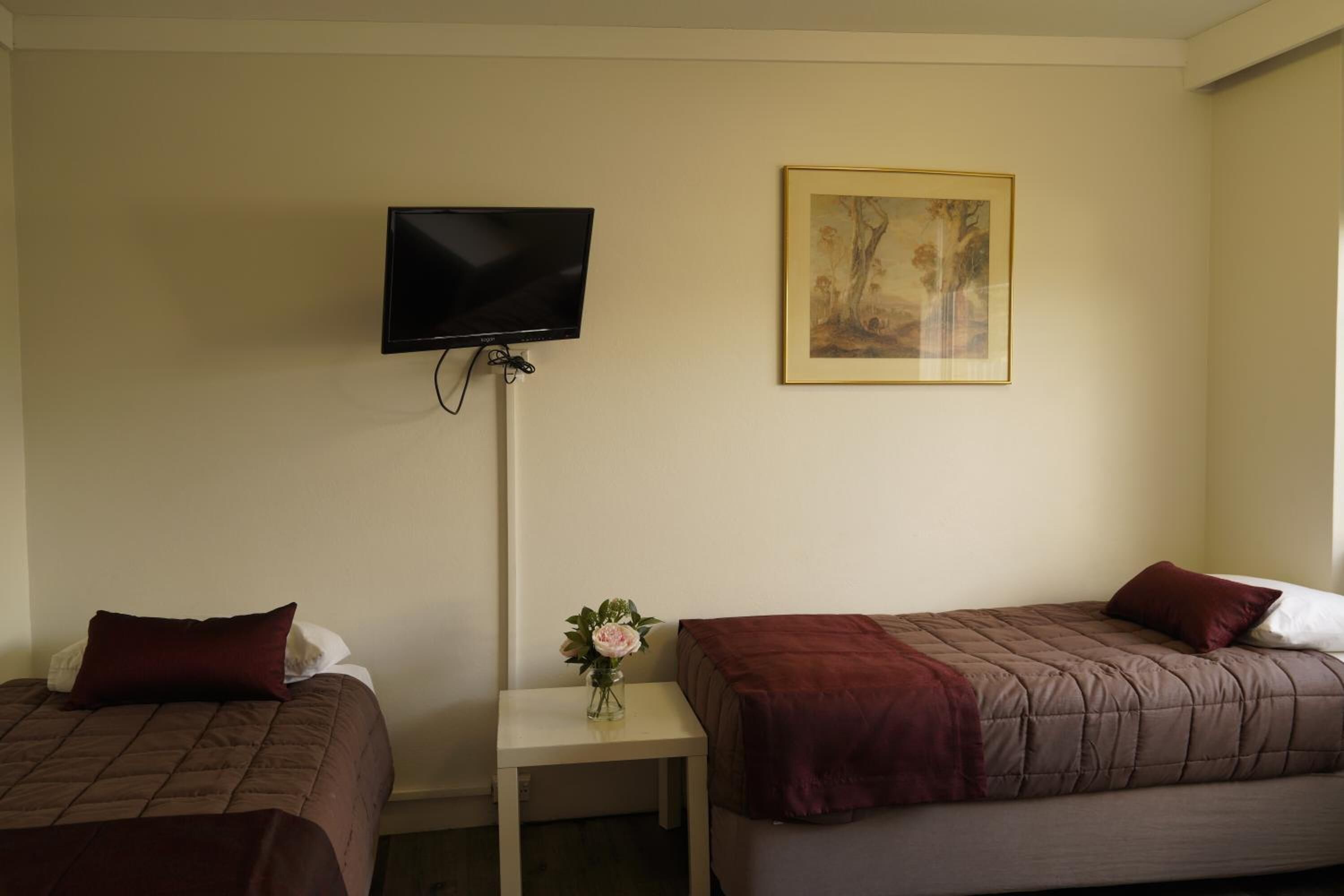 A hotel room with two beds, a TV mounted on the wall, a small table with a vase of flowers, and a framed picture on the wall.