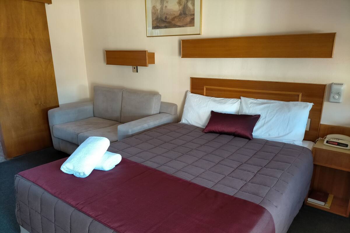 A hotel room with a bed, a sofa, a bedside table with a phone, and a wall-mounted shelf.