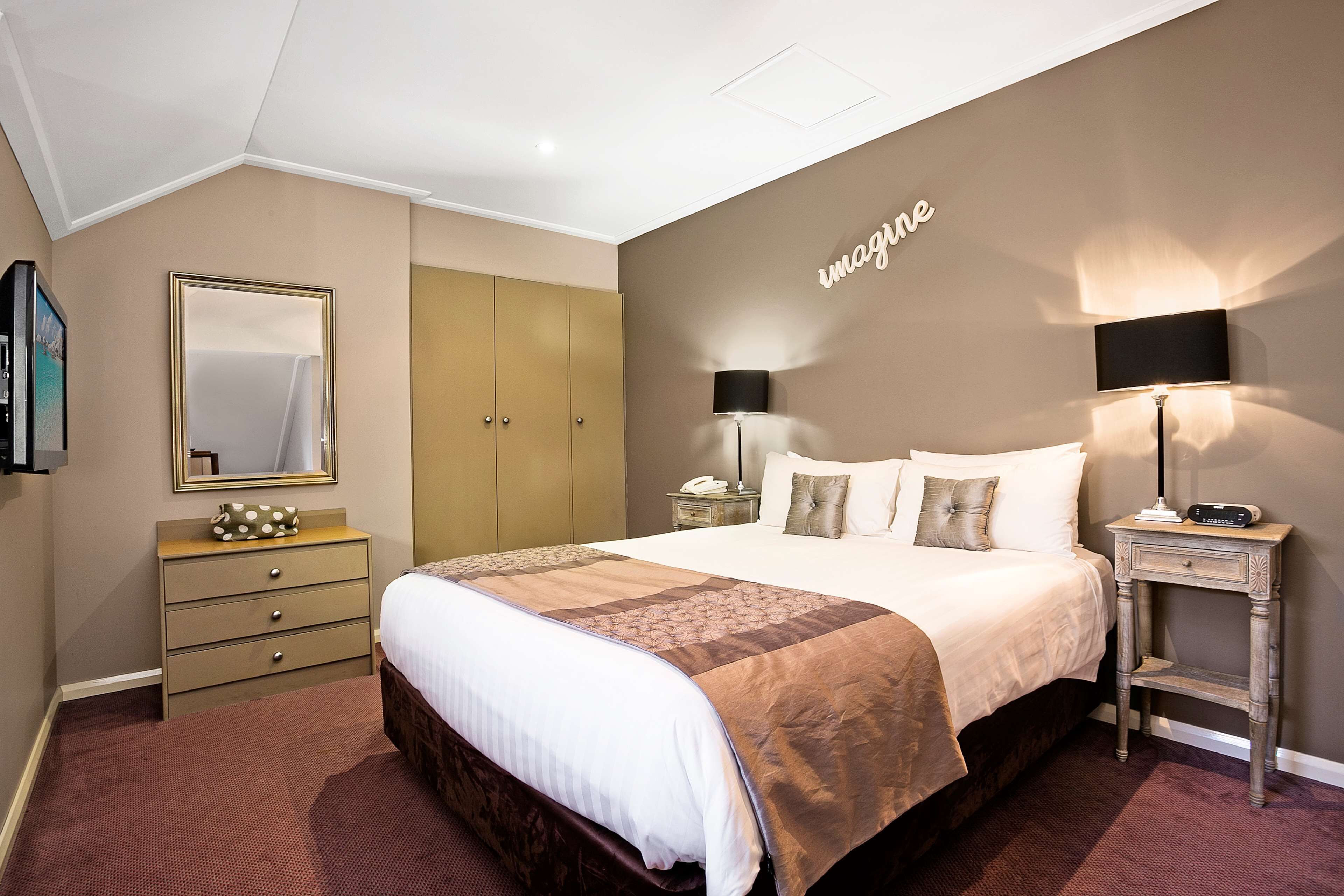 A hotel room with a large bed, a dresser, a nightstand, a lamp, and a mirror on the wall.