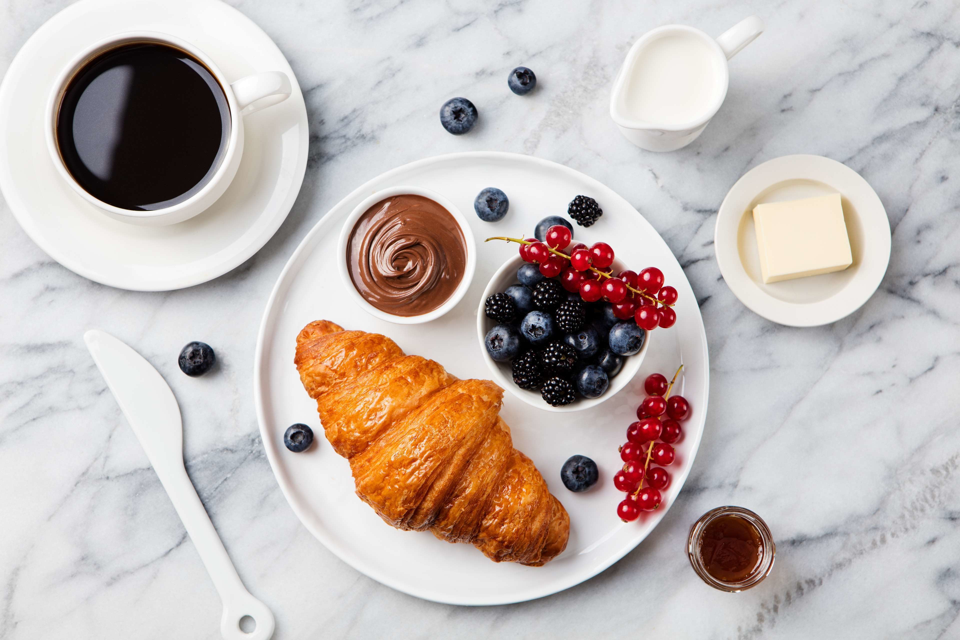 Una mesa de mármol con un croissant, crema de chocolate, arándanos, zarzamoras y un vaso de leche.