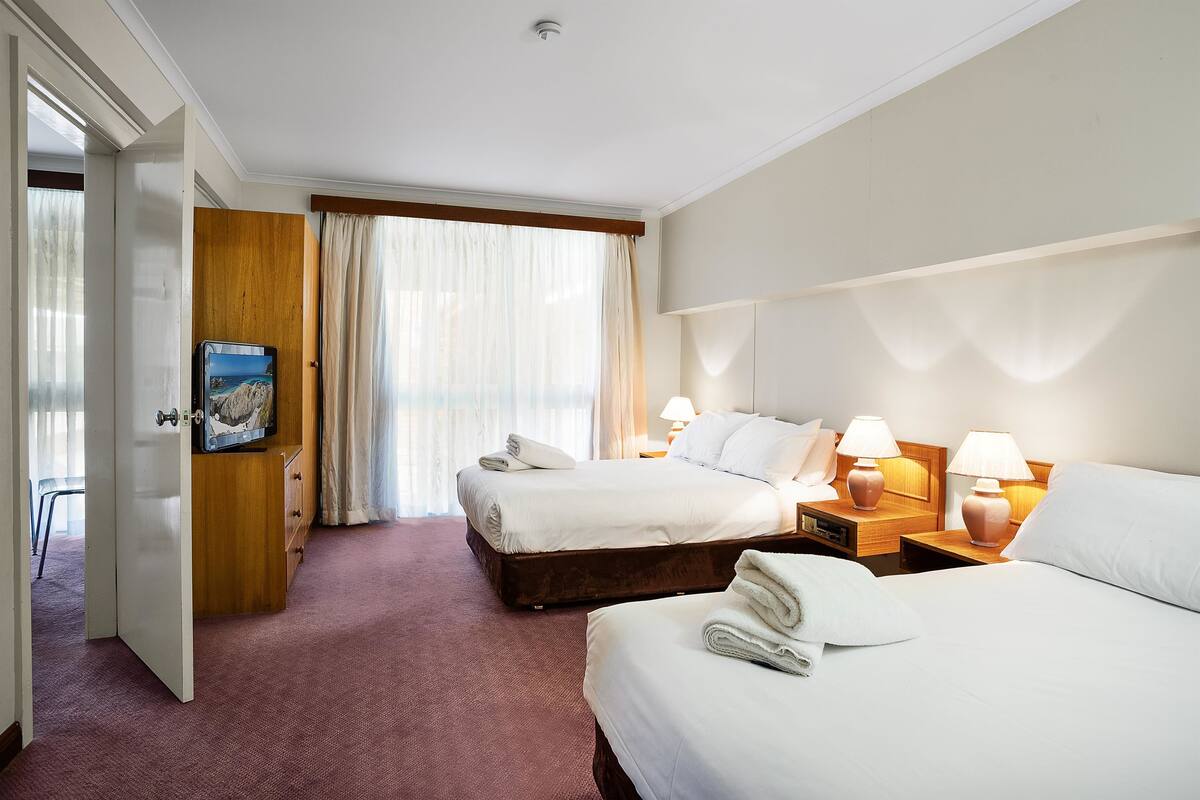 A hotel room with two beds, a wooden wardrobe, a desk with lamps, and a window with curtains.