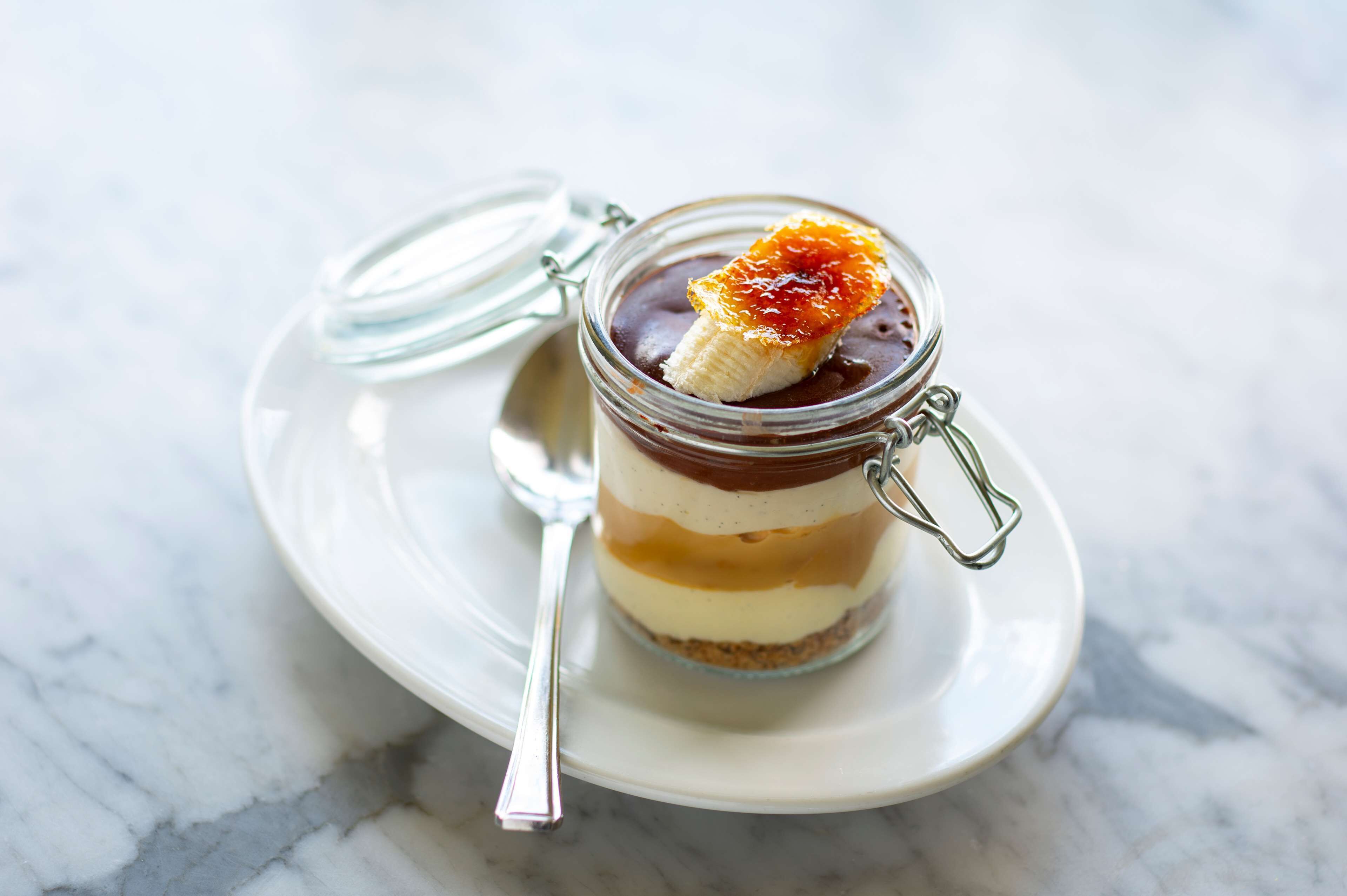 A layered dessert in a glass jar on a white plate with a spoon.