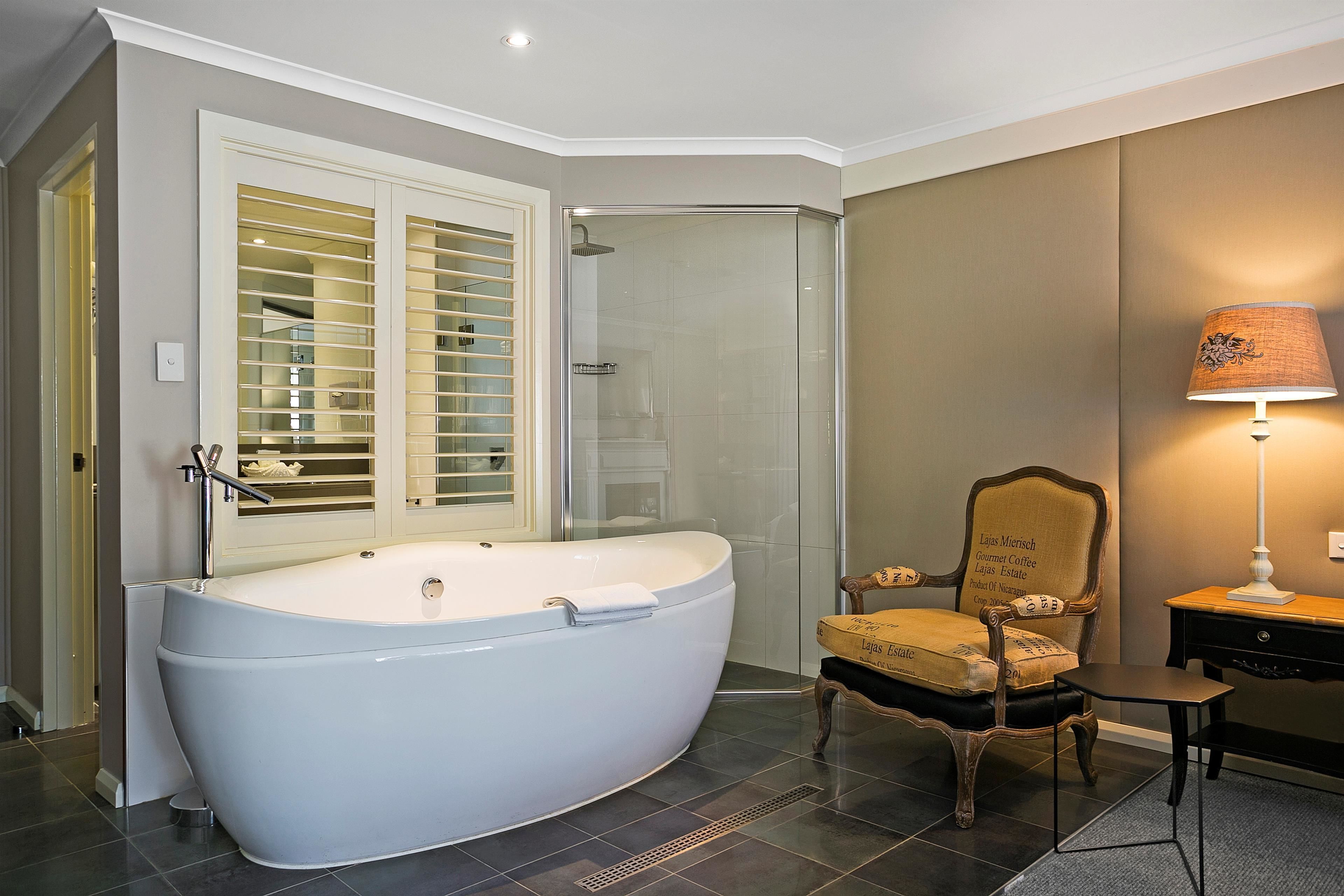 A modern bathroom with a freestanding bathtub, a glass shower enclosure, a vanity with a lamp, and a chair.