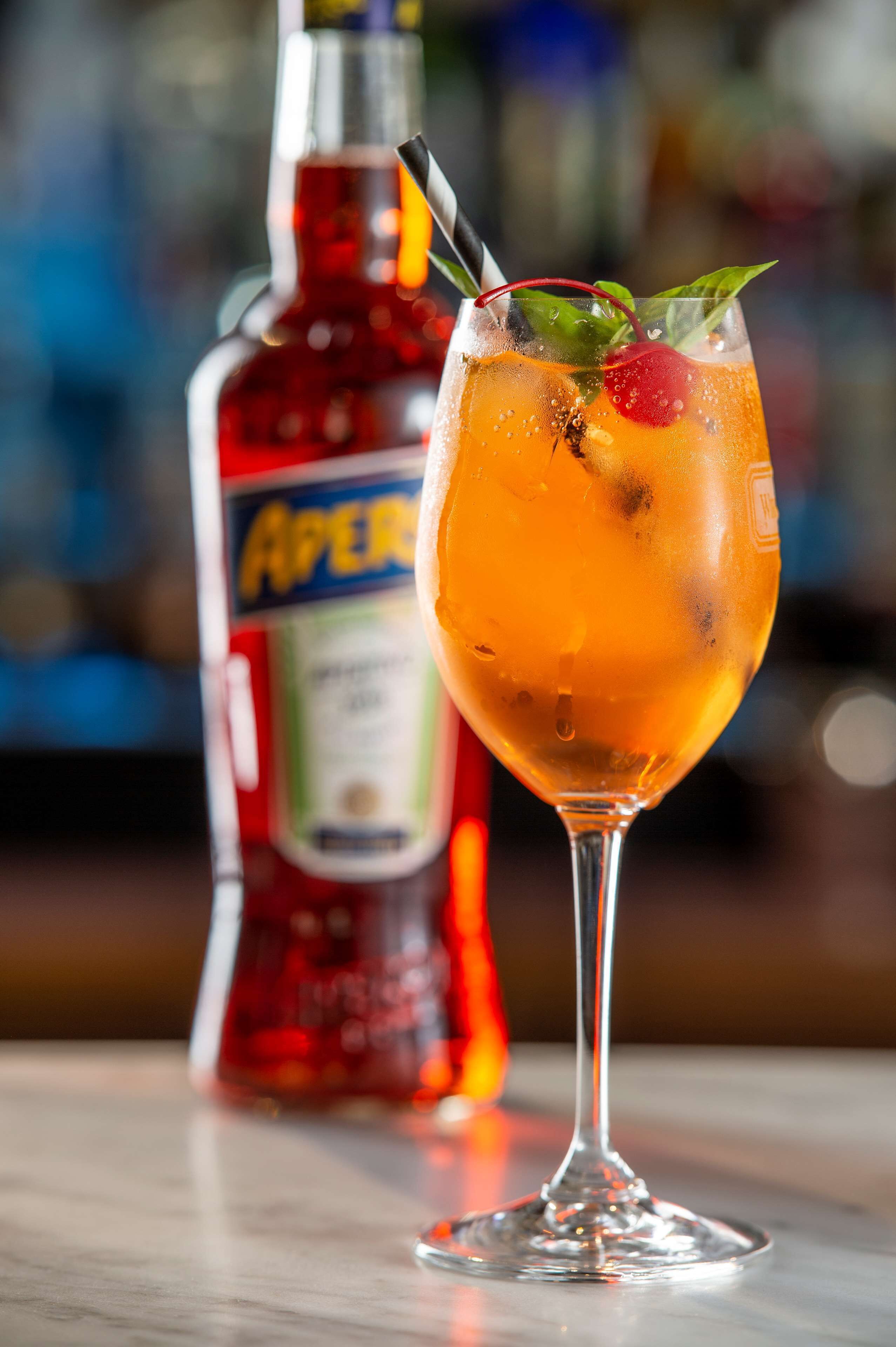 A cocktail in a stemmed glass with a cherry and mint garnish, and a bottle of Aperol in the background.