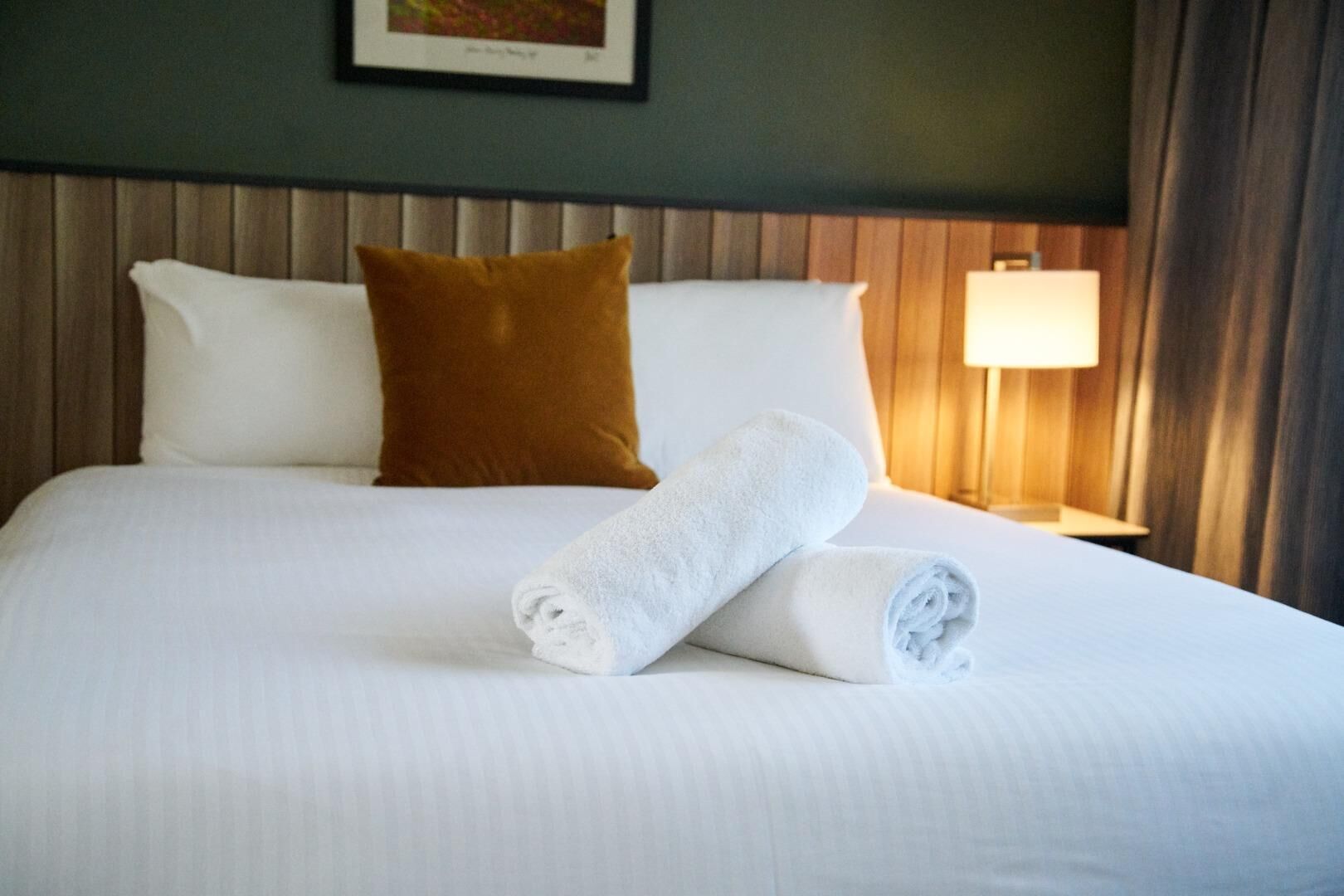 A neatly made bed with white linens, a brown pillow, and a rolled white towel.