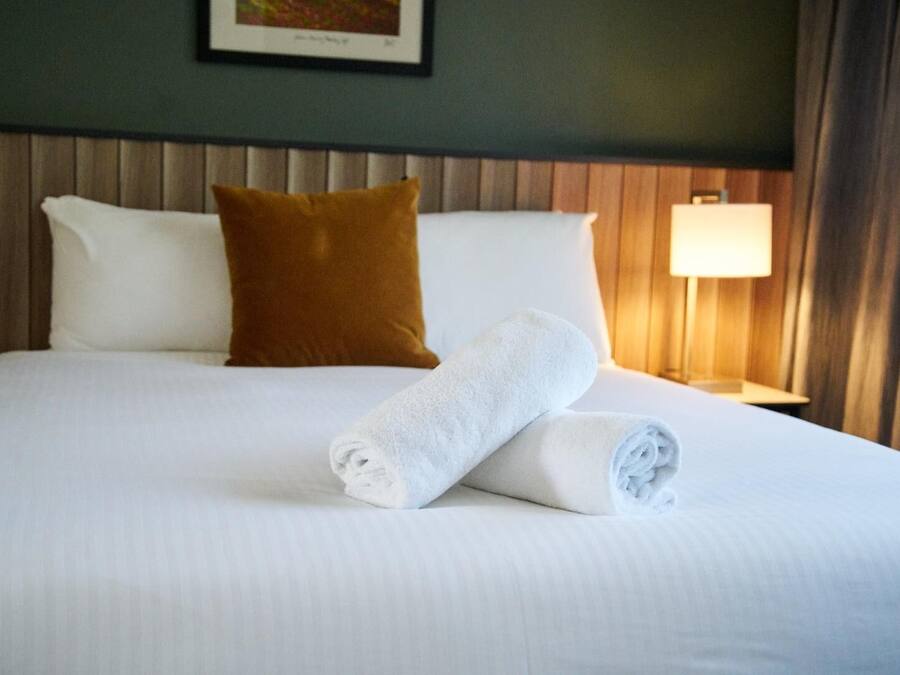A neatly made bed with white linens, a brown pillow, and a rolled white towel.