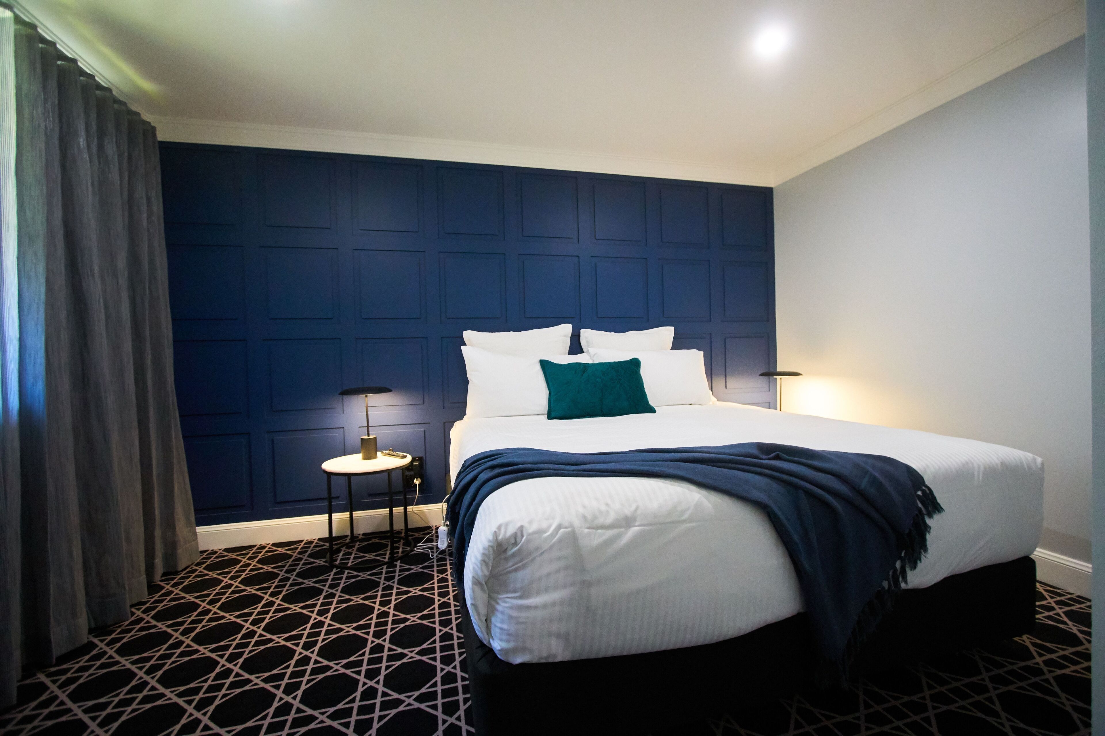 A hotel room with a large bed, dark blue wall paneling, a patterned floor, and a small bedside table with a lamp.