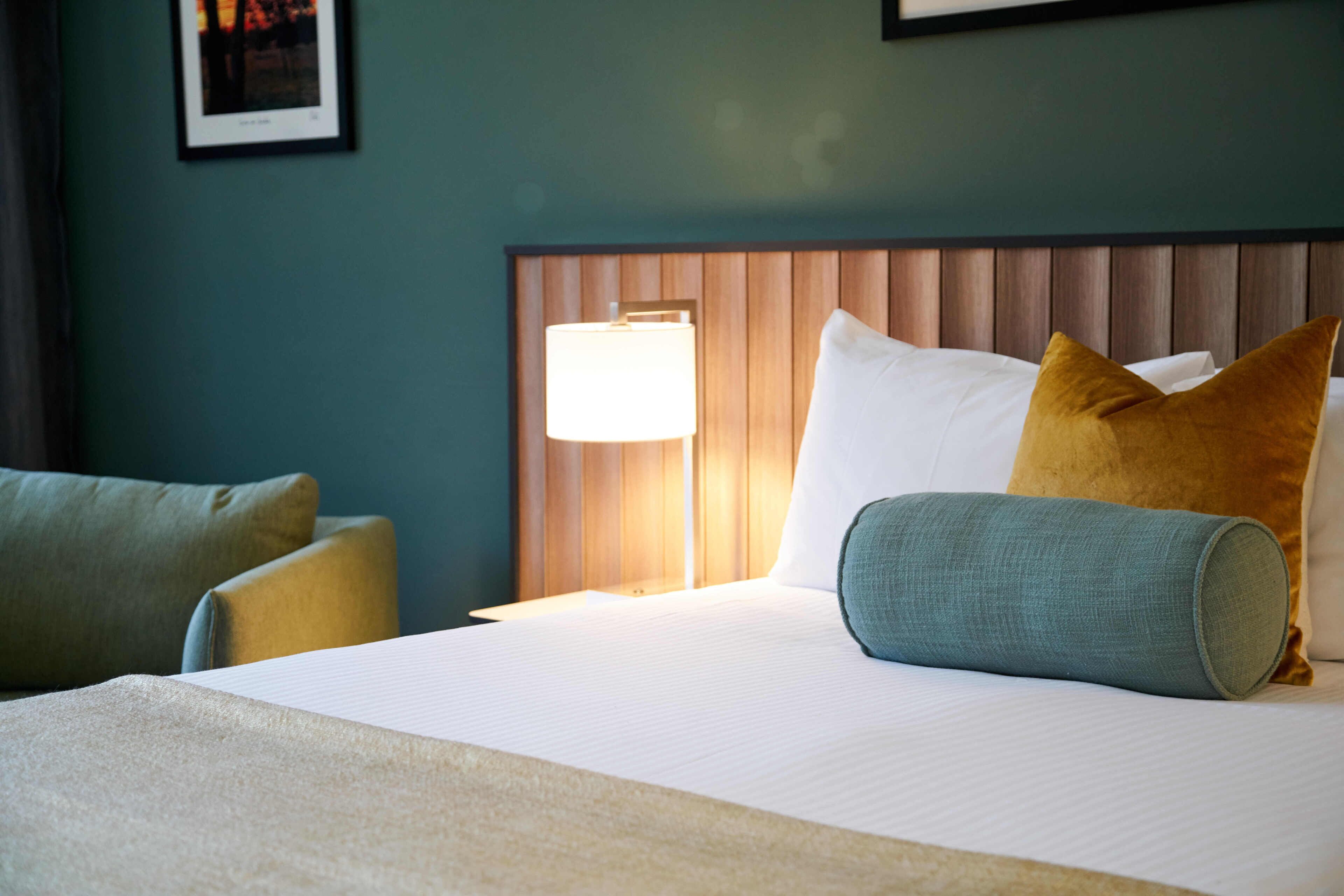 A hotel room with a bed, pillows, a lamp, and a framed picture on the wall.