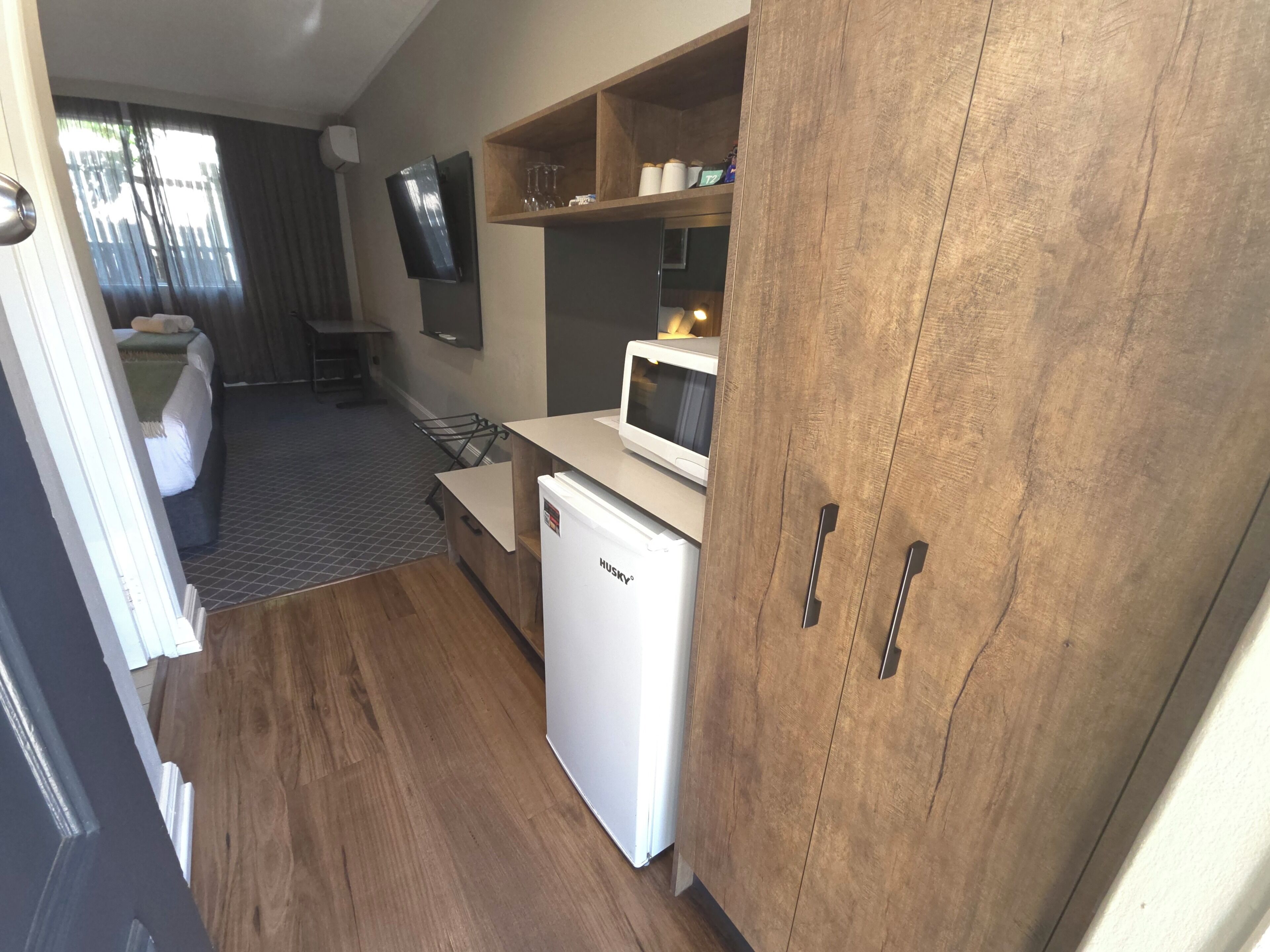 A compact kitchen with a microwave, a small refrigerator, wooden cabinets, and a shelf with glassware.