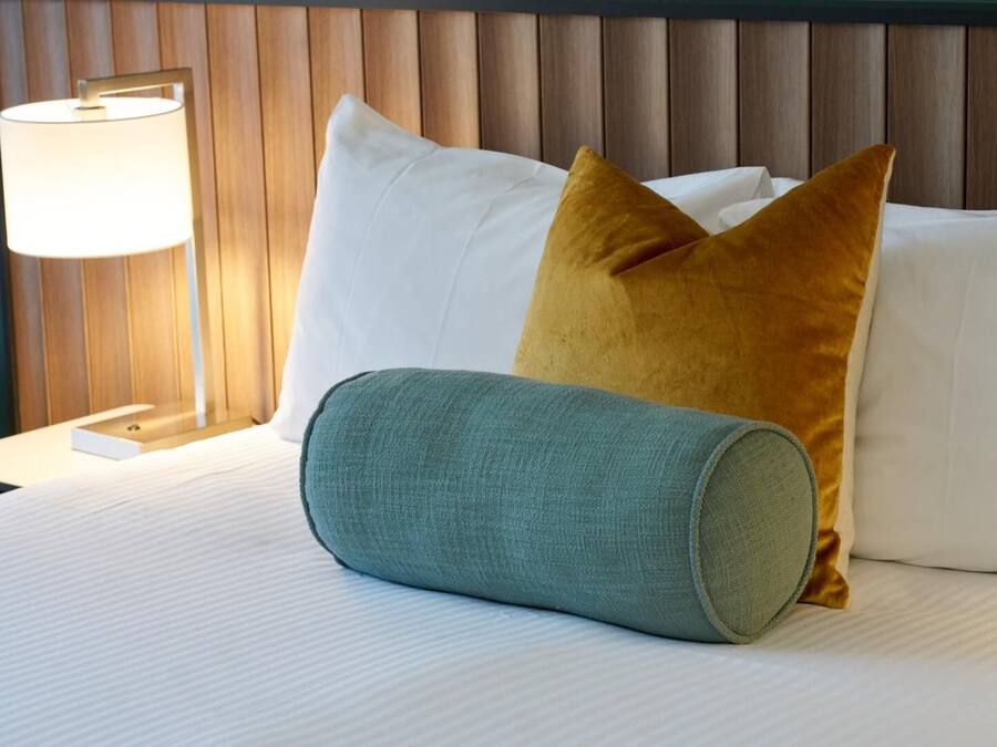 A neatly made bed with white linens, a teal cylindrical pillow, and two mustard-colored pillows. A bedside lamp with a white shade is visible on the left.