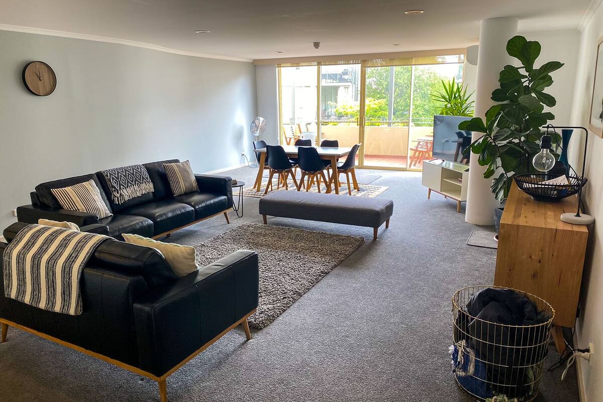 Uma sala de estar moderna com um sofá de couro preto, um banquinho cinza e uma área de jantar com mesa e cadeiras. Há uma janela grande que oferece luz natural e vista para o exterior.