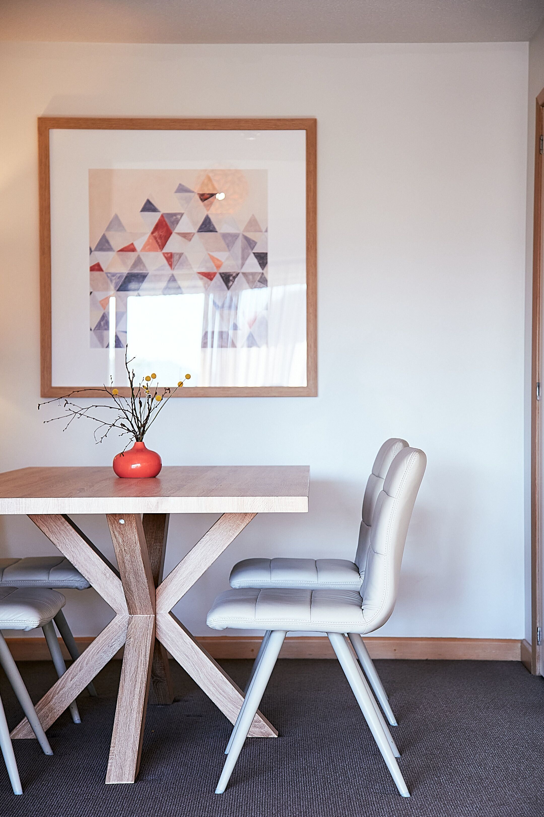 A dining area with a wooden table, two chairs, a vase with branches, and a framed abstract painting on the wall.