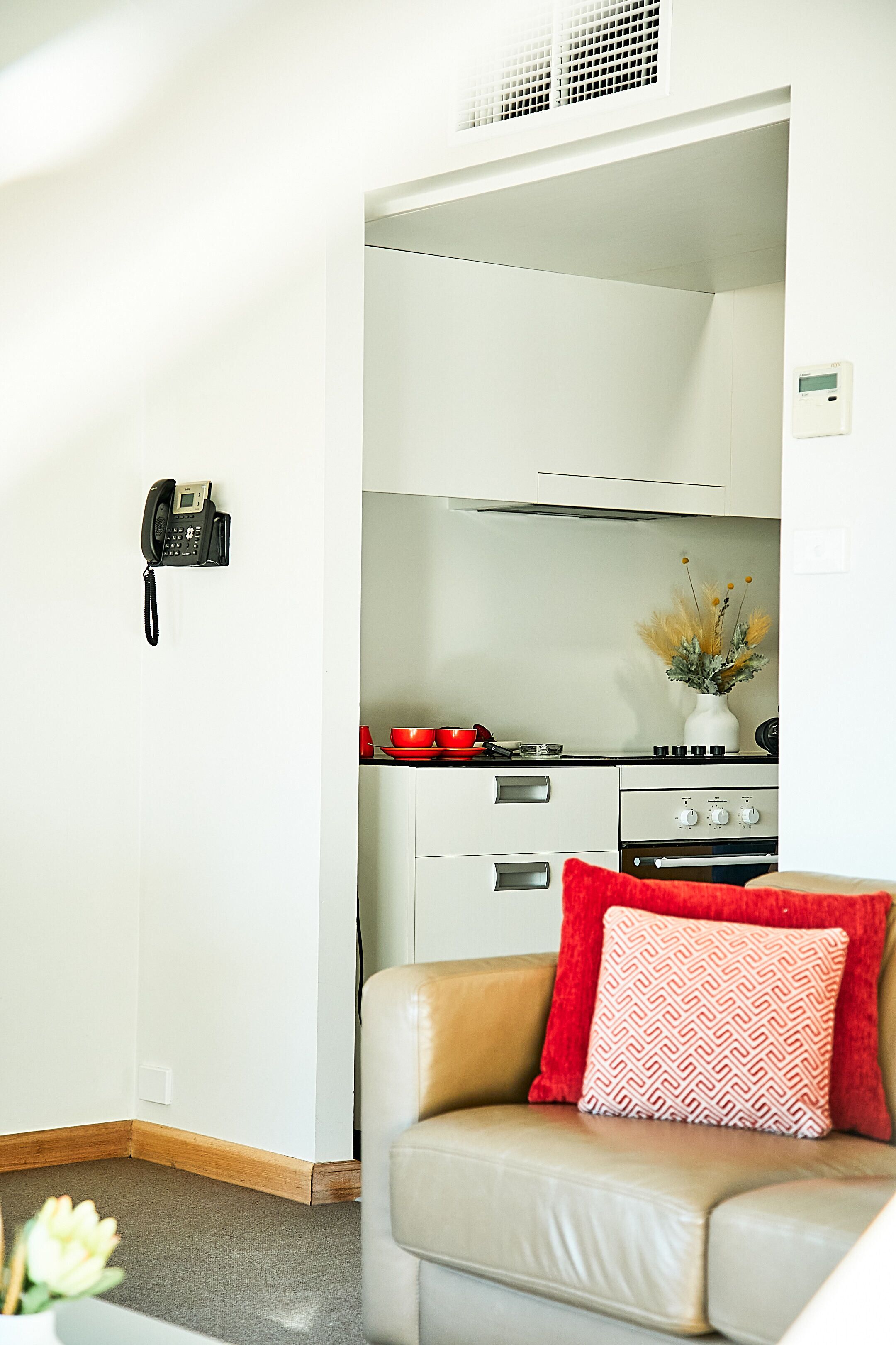 A modern kitchen with white cabinets, a stove, and a red pot. There is a phone mounted on the wall and a vase with dried flowers on the counter.