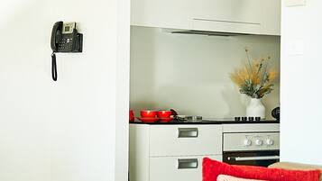 A modern kitchen with white cabinets, a stove, and a red pot. There is a phone mounted on the wall and a vase with dried flowers on the counter.