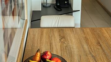 Una mesa de madera con un frutero, una silla blanca, un teléfono y una estantería con libros y una planta.