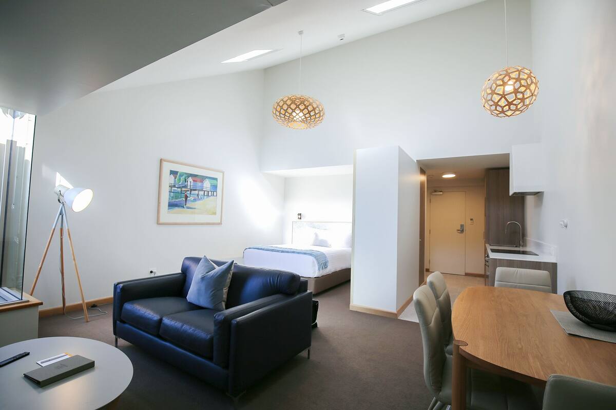 Quarto de hotel moderno com sofá, mesa de jantar, cama e cozinha compacta.