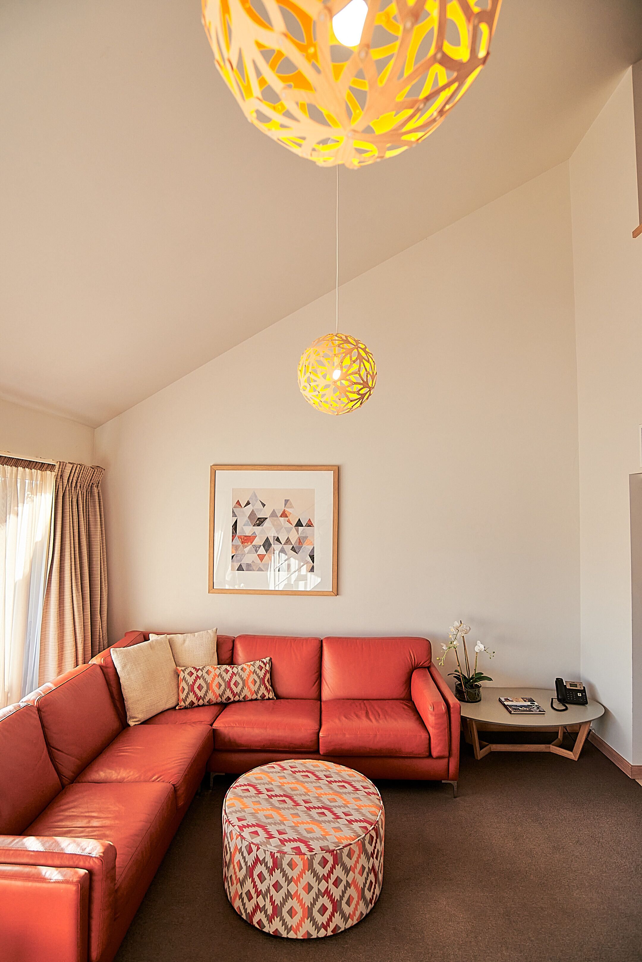 A living room with a sectional sofa, a patterned ottoman, a small table with a phone, and a hanging light fixture.