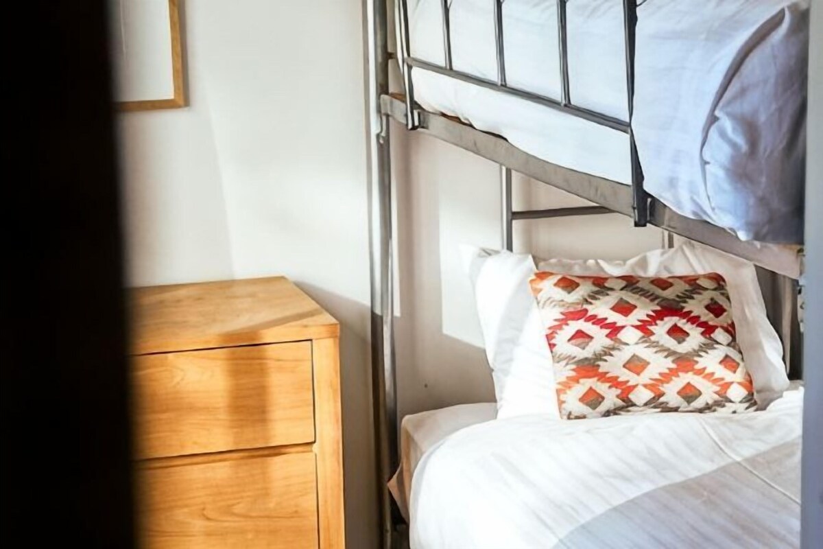 Una cama litera con un cojín estampado, una mesita de noche de madera y un cuadro en la pared.