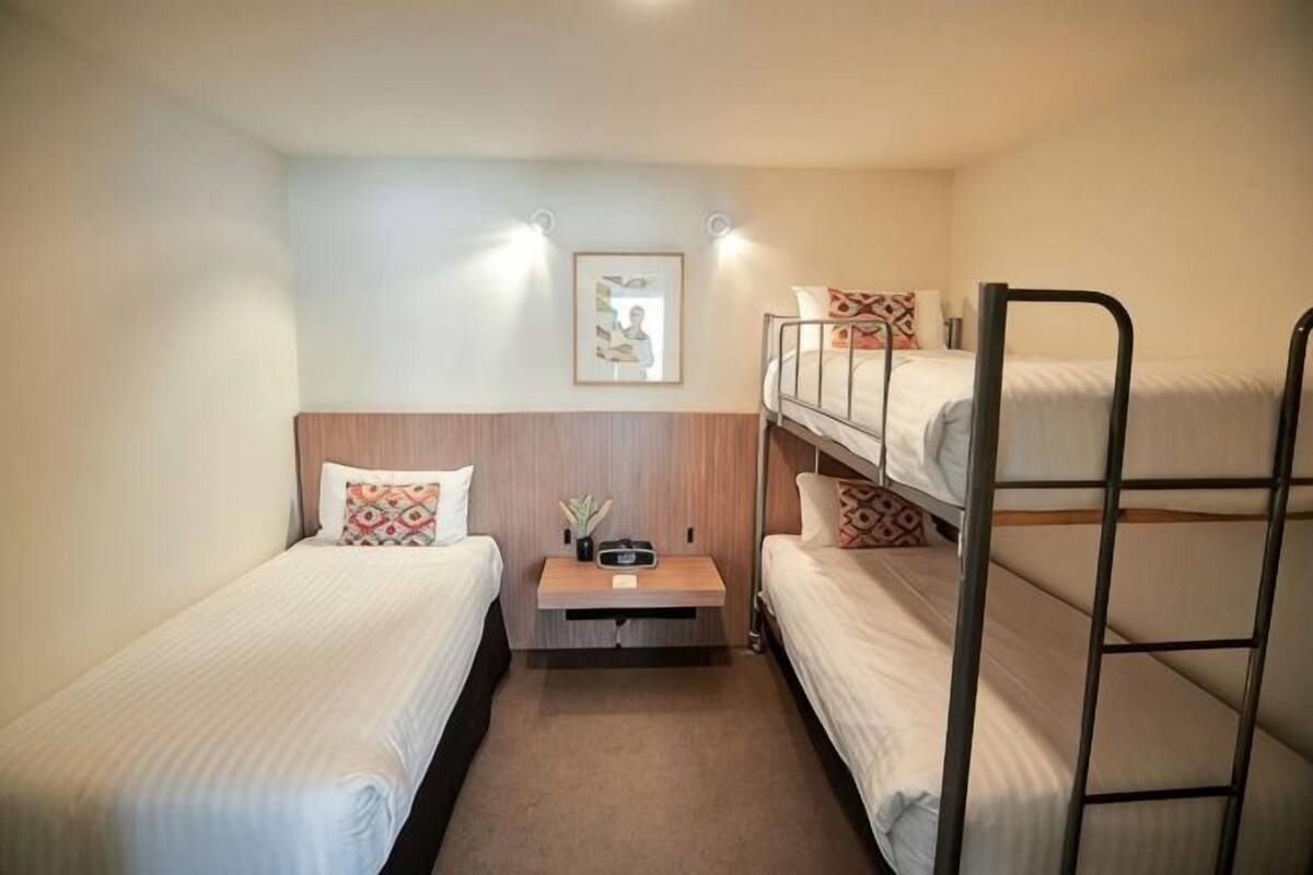 Un pequeño hotel con una sola habitación, equipado con literas, una mesita de noche con lámpara y un cuadro en la pared.