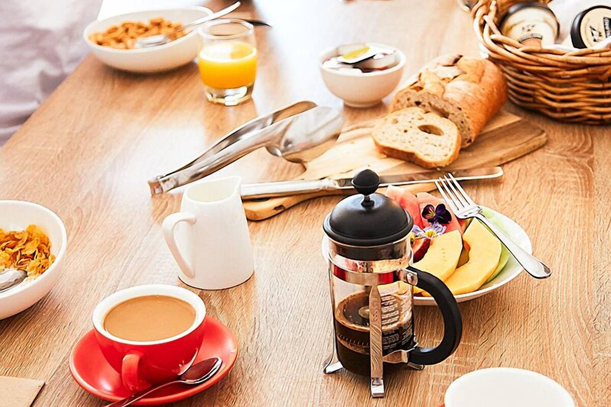 Mesa de café da manhã com diversos alimentos, uma cafeteira e um smartphone em uso.