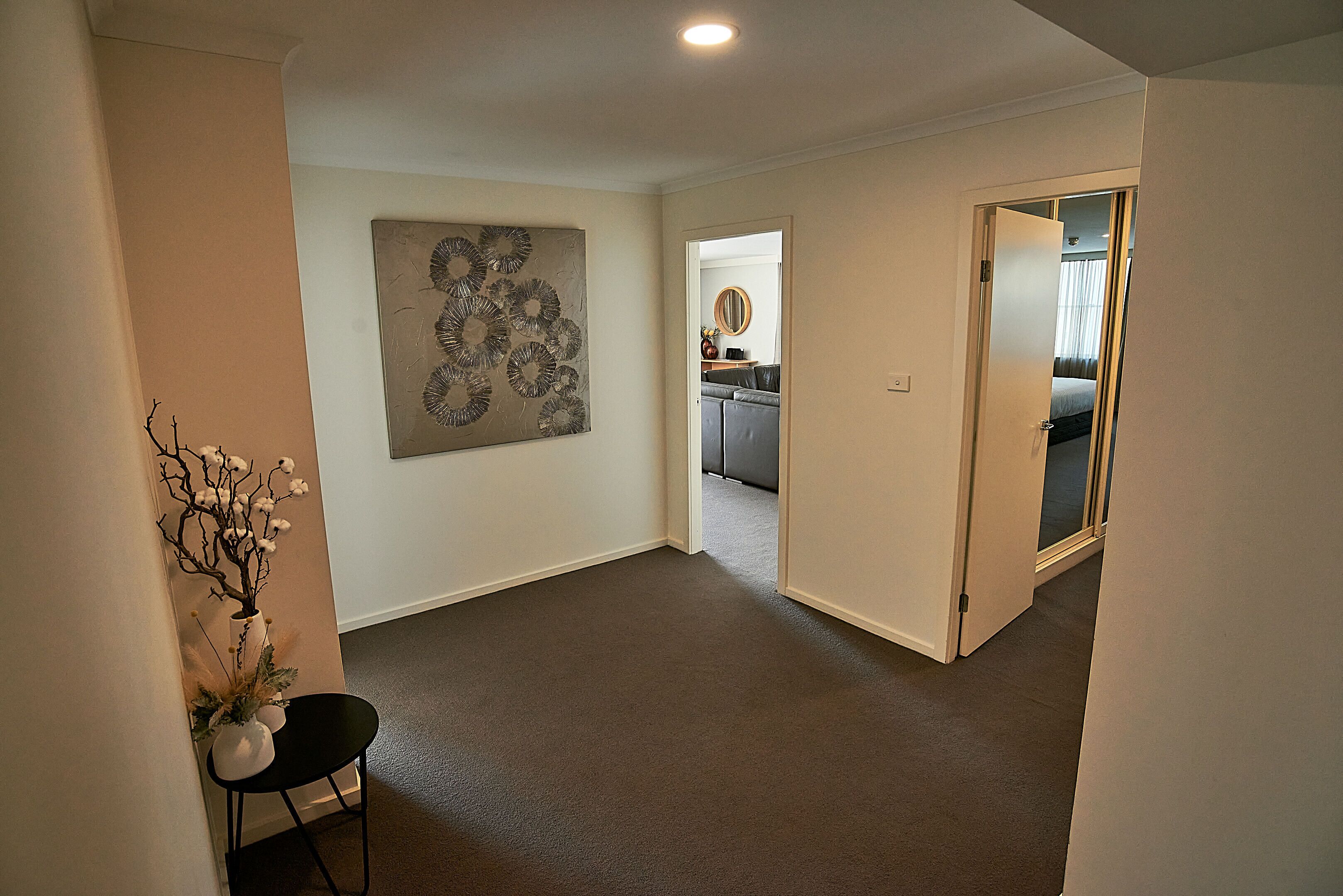 A hallway with a carpeted floor, a white vase with branches on a black chair, and a wall-mounted abstract art piece.