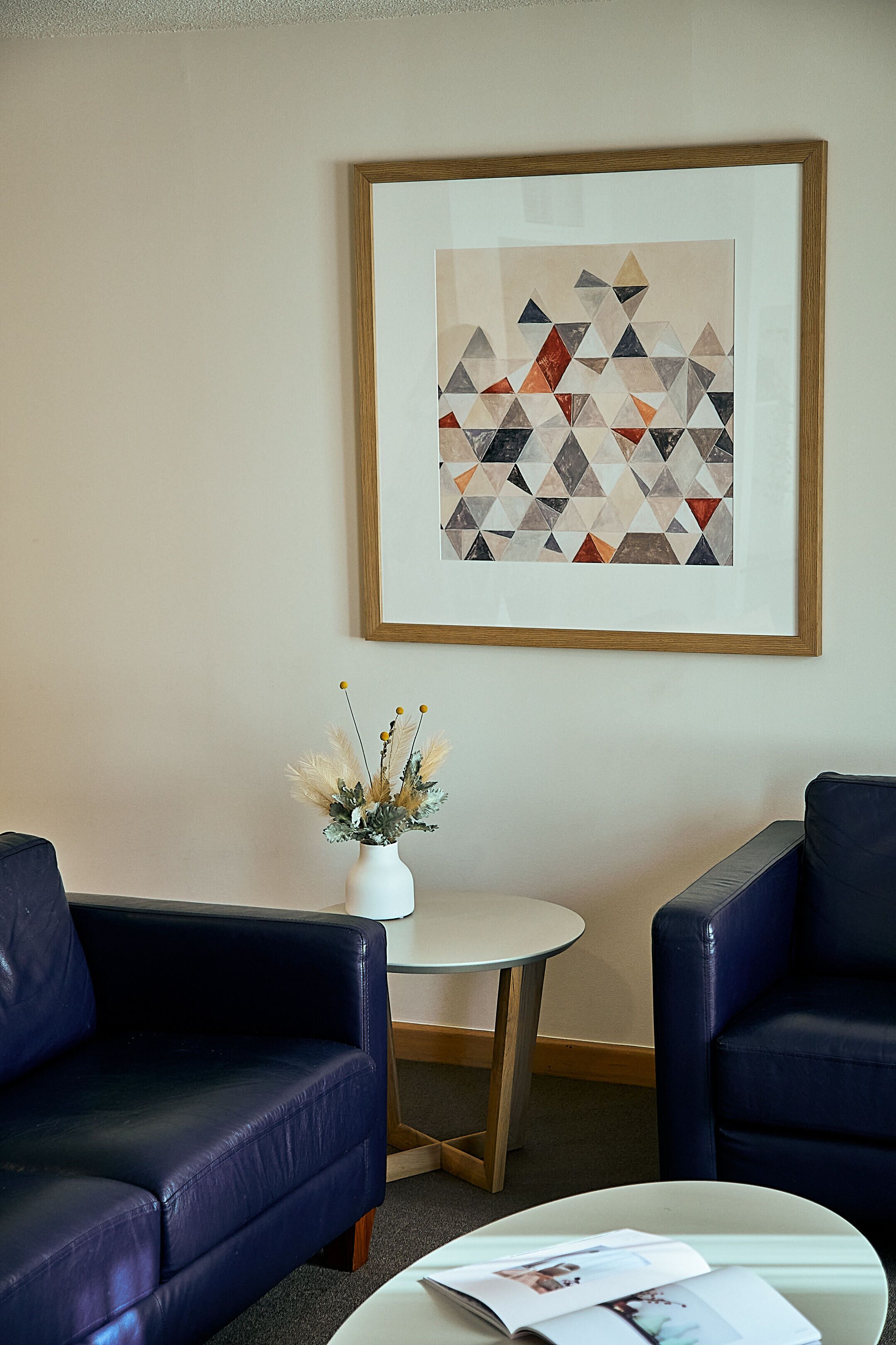 Una habitación con un cuadro abstracto enmarcado, una mesita blanca, dos sillones de cuero azul y un florero con plantas secas.