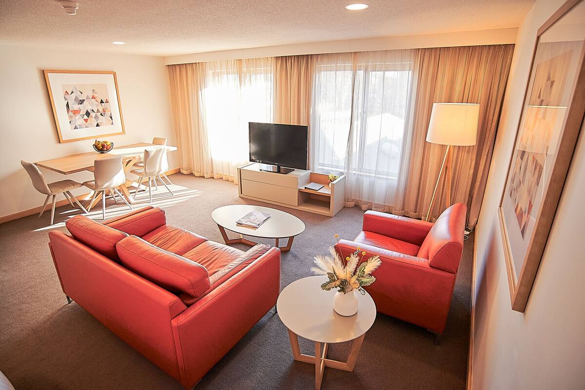 Quarto de hotel moderno com sofá vermelho, televisão de tela plana, mesa de jantar com cadeiras e pequena mesa de centro.