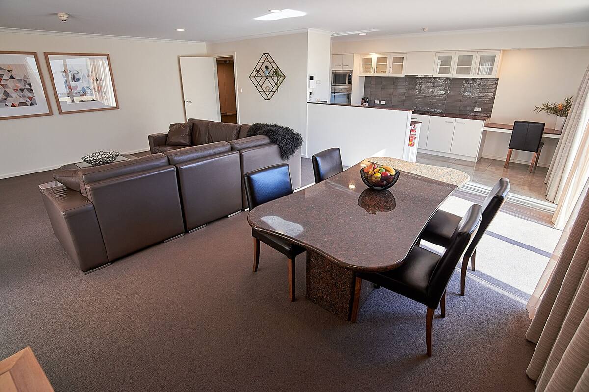 Uma sala de estar moderna com um sofá de couro marrom, uma mesa de jantar com cadeiras e uma cozinha ao fundo.