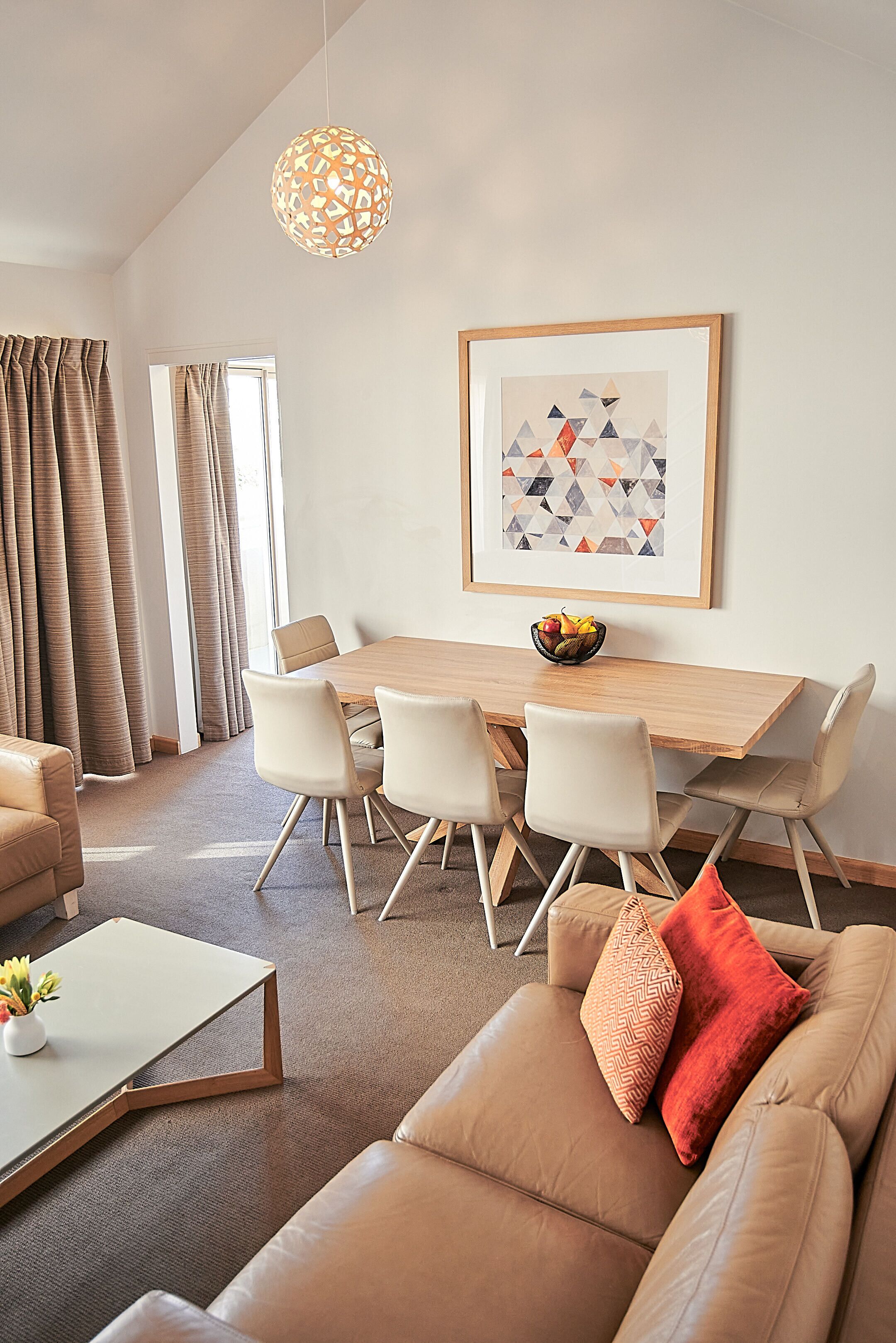 A modern living room with a dining area, featuring a wooden table, white chairs, a beige sofa, and a hanging light fixture.