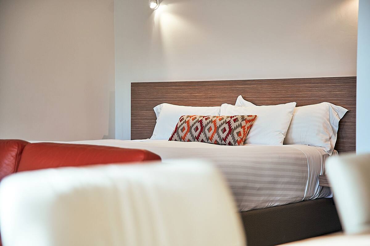 Cama bem arrumada com roupa de cama branca e listrada, almofada estampada e cabeceira de madeira.