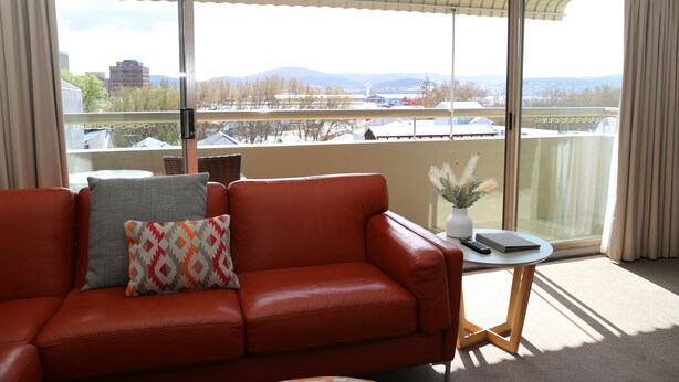 Une pièce avec un canapé en cuir rouge, une petite table ronde sur laquelle se trouve une télécommande, et une vue sur le paysage urbain à travers de grandes portes vitrées.
