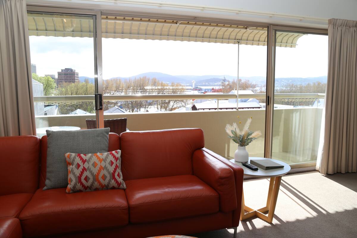 Quarto com sofá de couro vermelho, uma pequena mesa redonda com um controle remoto e vista para um horizonte urbano através de grandes portas de vidro.