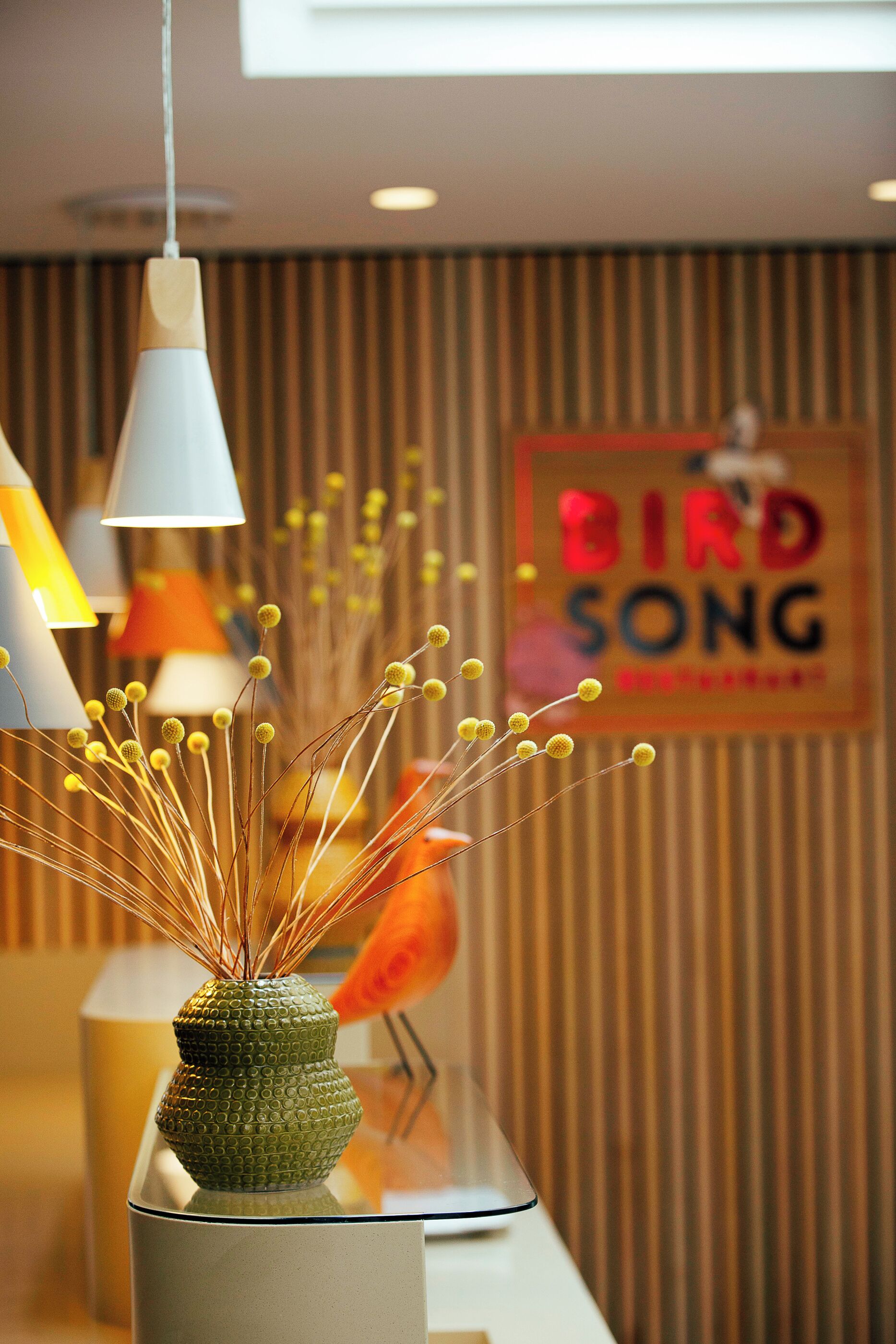 Ein Empfangstresen mit einer grünen Vase und dekorativen Zweigen, ein Schild mit der Aufschrift 'BIRD SONG' und eine gestreifte Wand im Hintergrund.