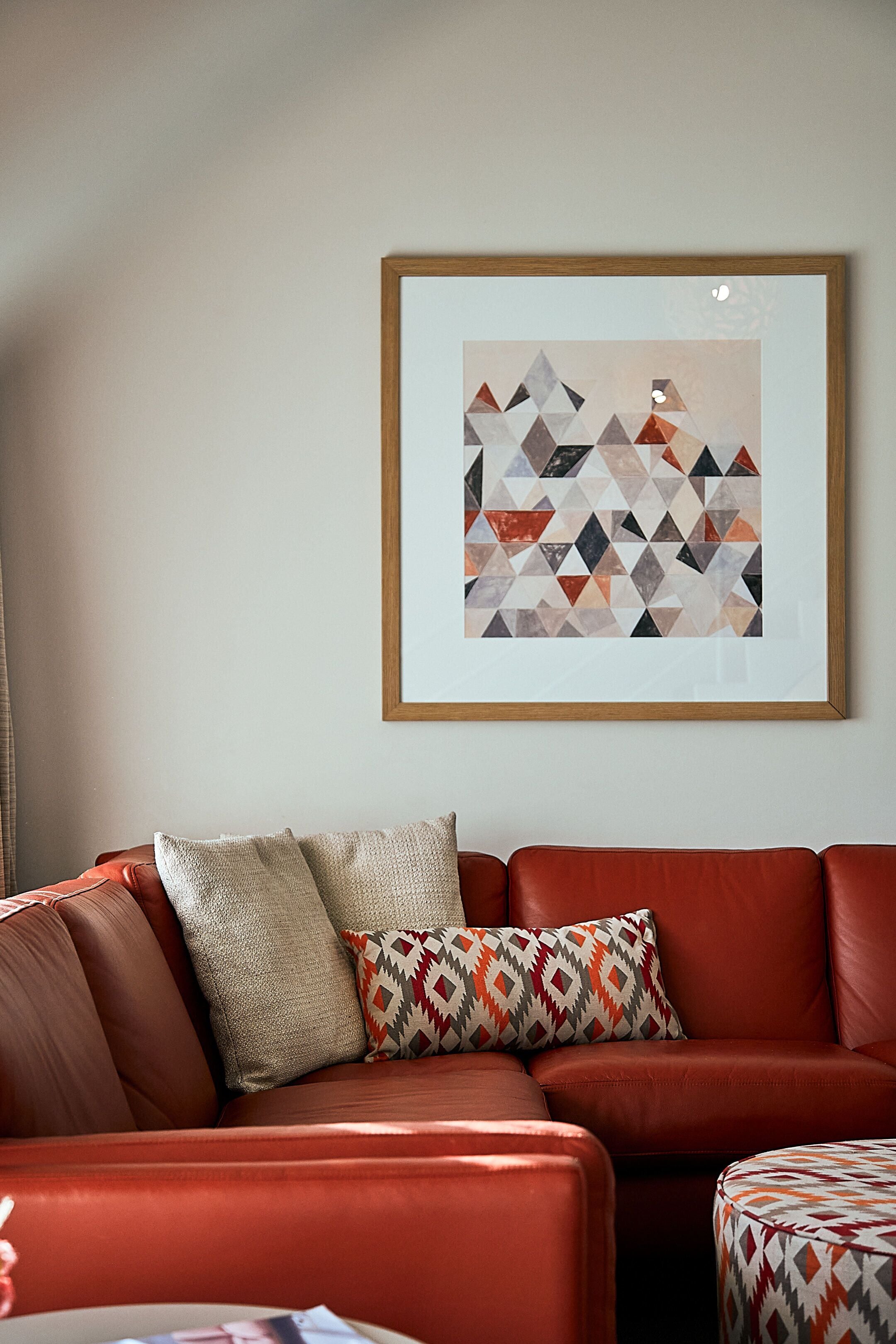 A red leather sofa with patterned cushions, a framed abstract artwork on the wall, and a small ottoman with a geometric pattern.