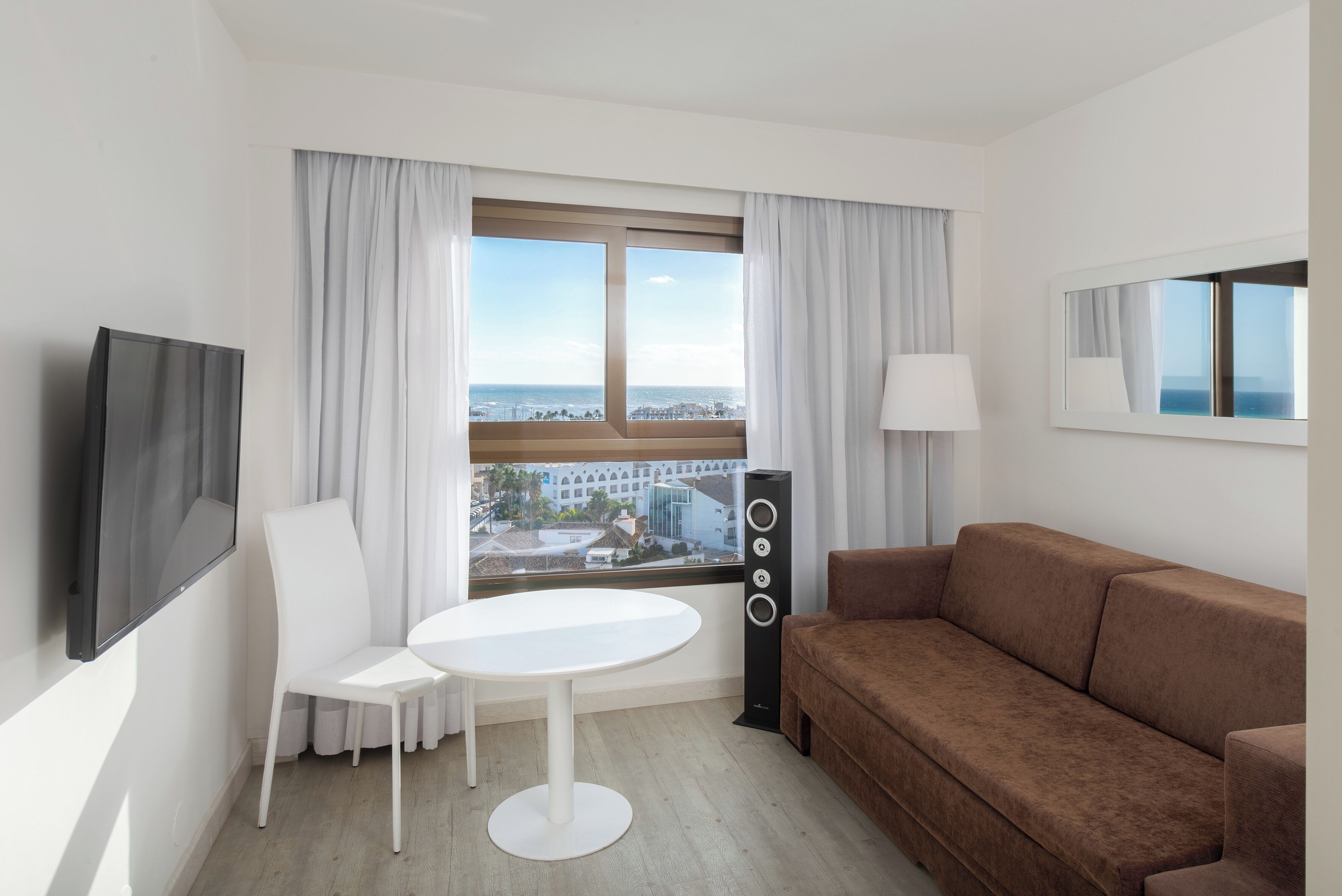 Ein modernes Hotelzimmer mit einem großen Fenster, einem weißen Tisch, einem braunen Sofa, einem Fernseher und einer Musikanlage.