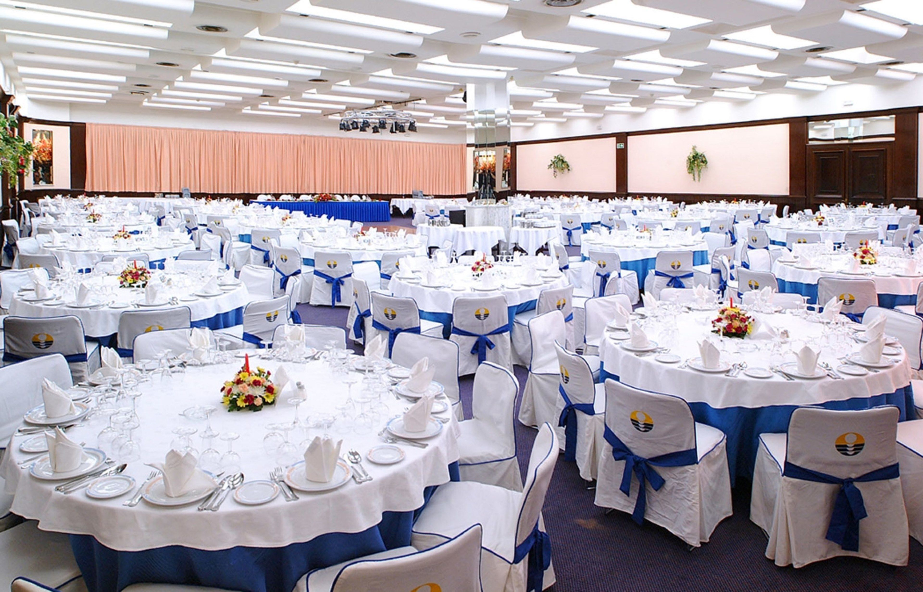 Un salón de banquetes con mesas redondas dispuestas para eventos, cada una con manteles blancos y fajas azules.