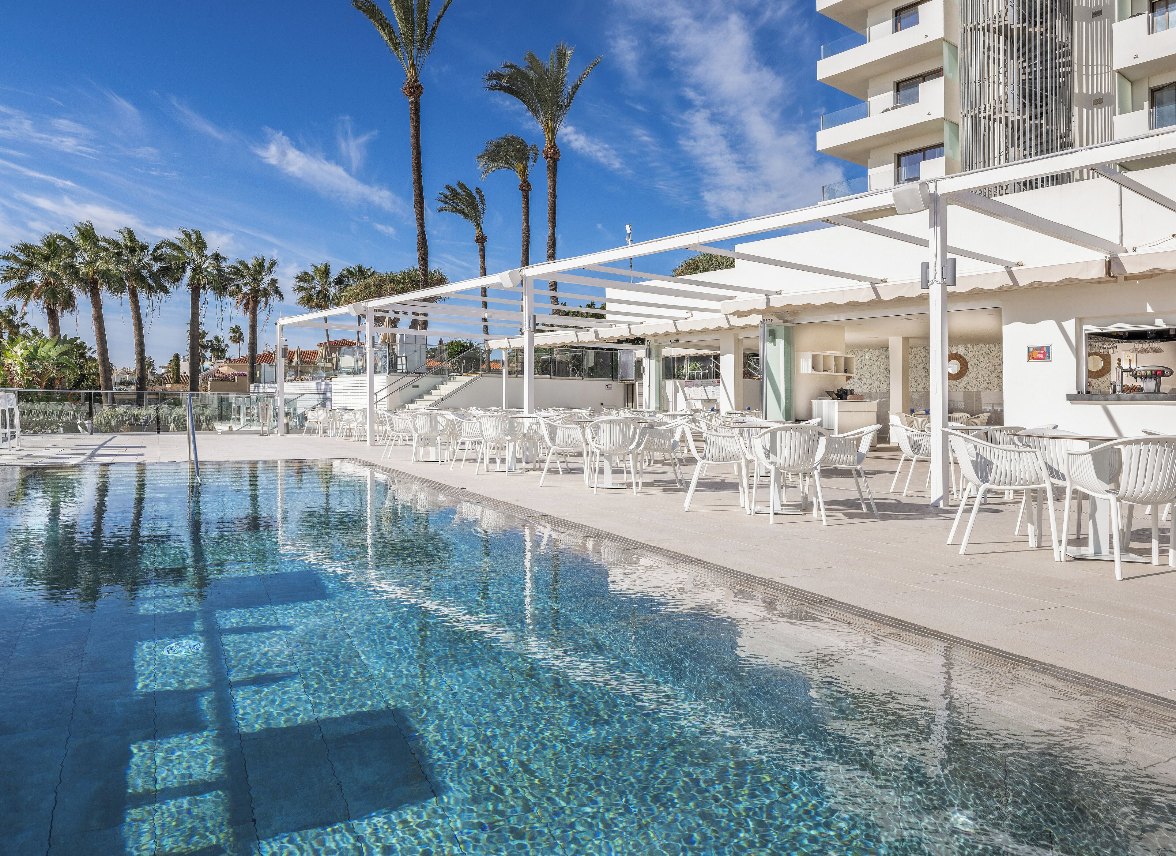 A pool area with clear water, white chairs, and palm trees.
