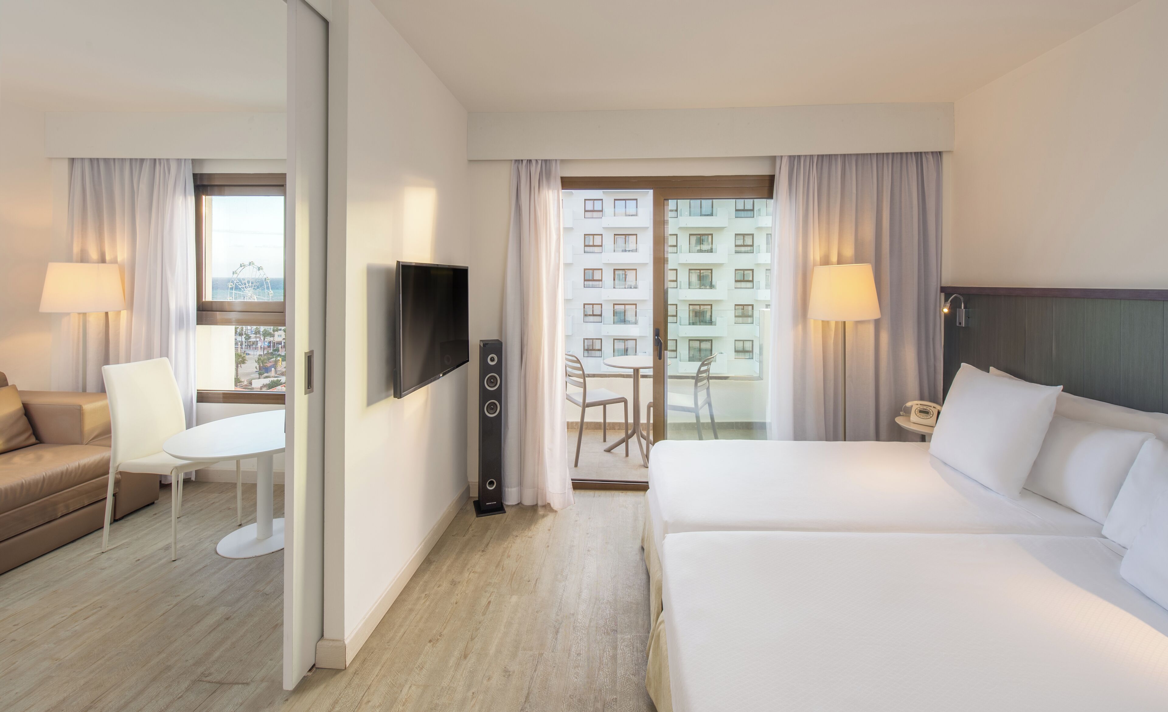 Habitación de hotel con cama, escritorio, silla, televisión y balcón con vista a edificios.