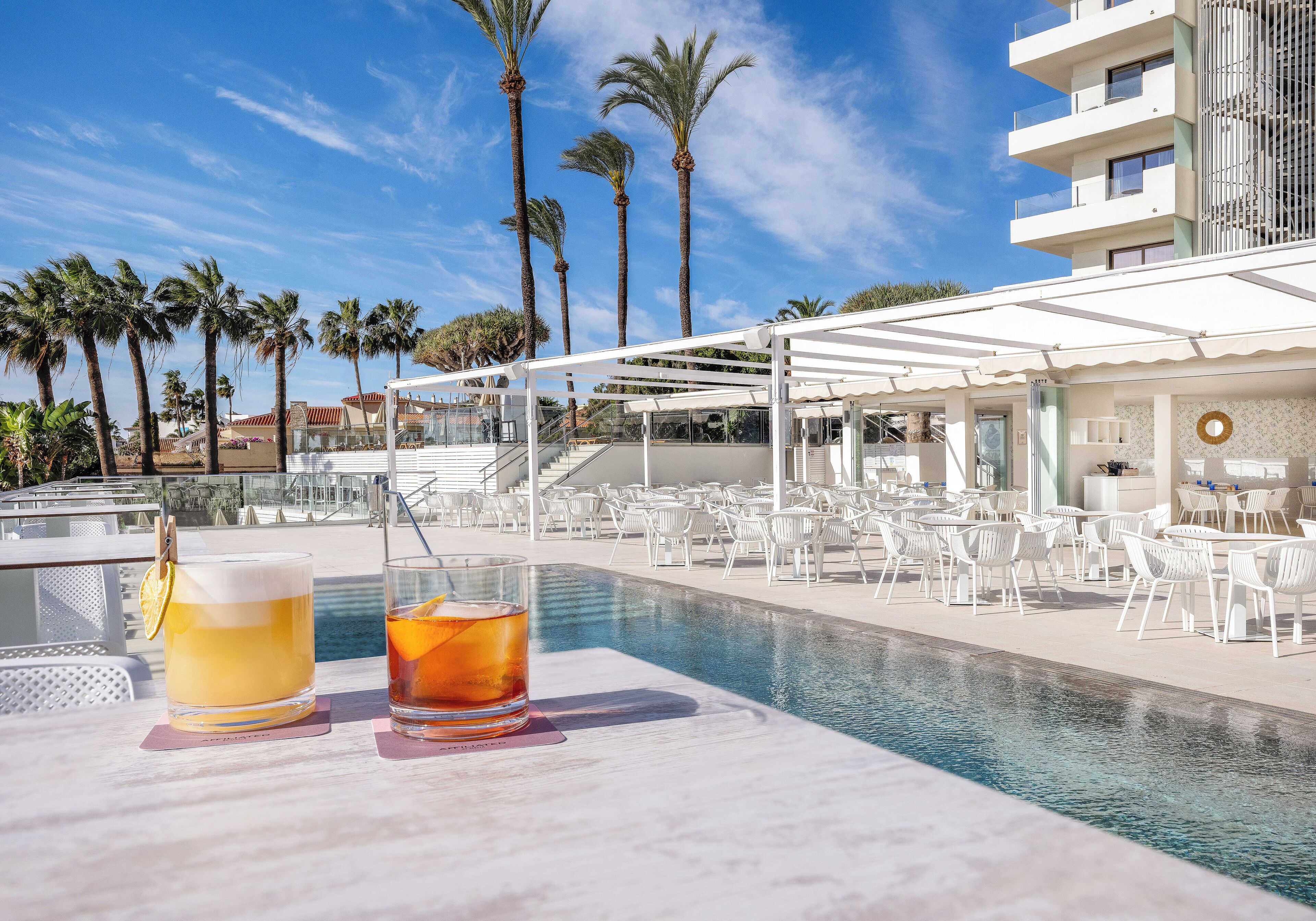 Zona junto a la piscina con dos bebidas sobre una mesa, rodeada de sillas blancas y un área de estar cubierta.