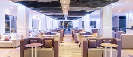 A modern hotel lobby with sofas and round tables, a patterned ceiling, and large windows.