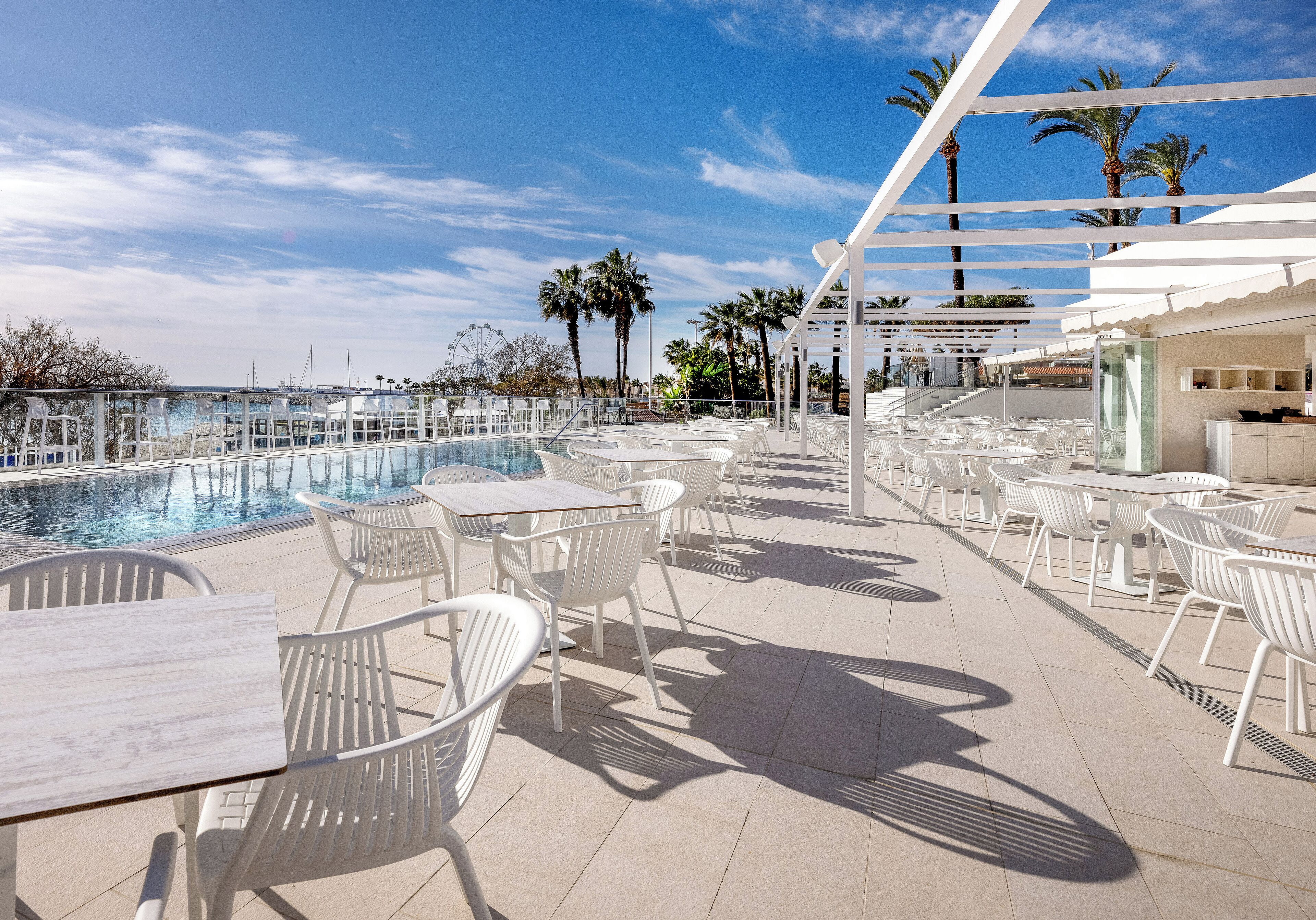 A poolside area with white chairs and tables, a clear sky, and palm trees in the background.