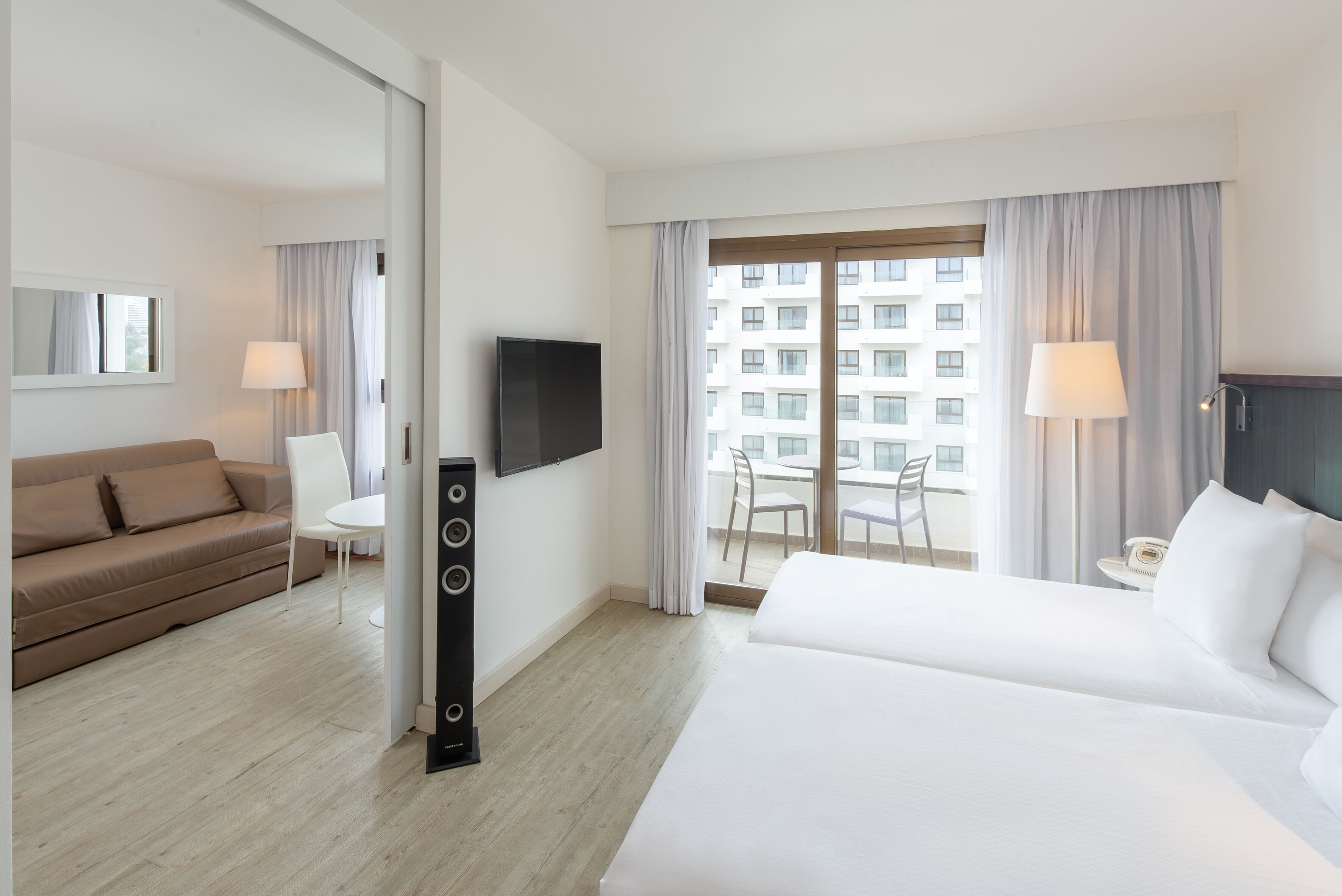 A hotel room with a bed, a sofa, a TV, a desk, and a balcony with curtains.