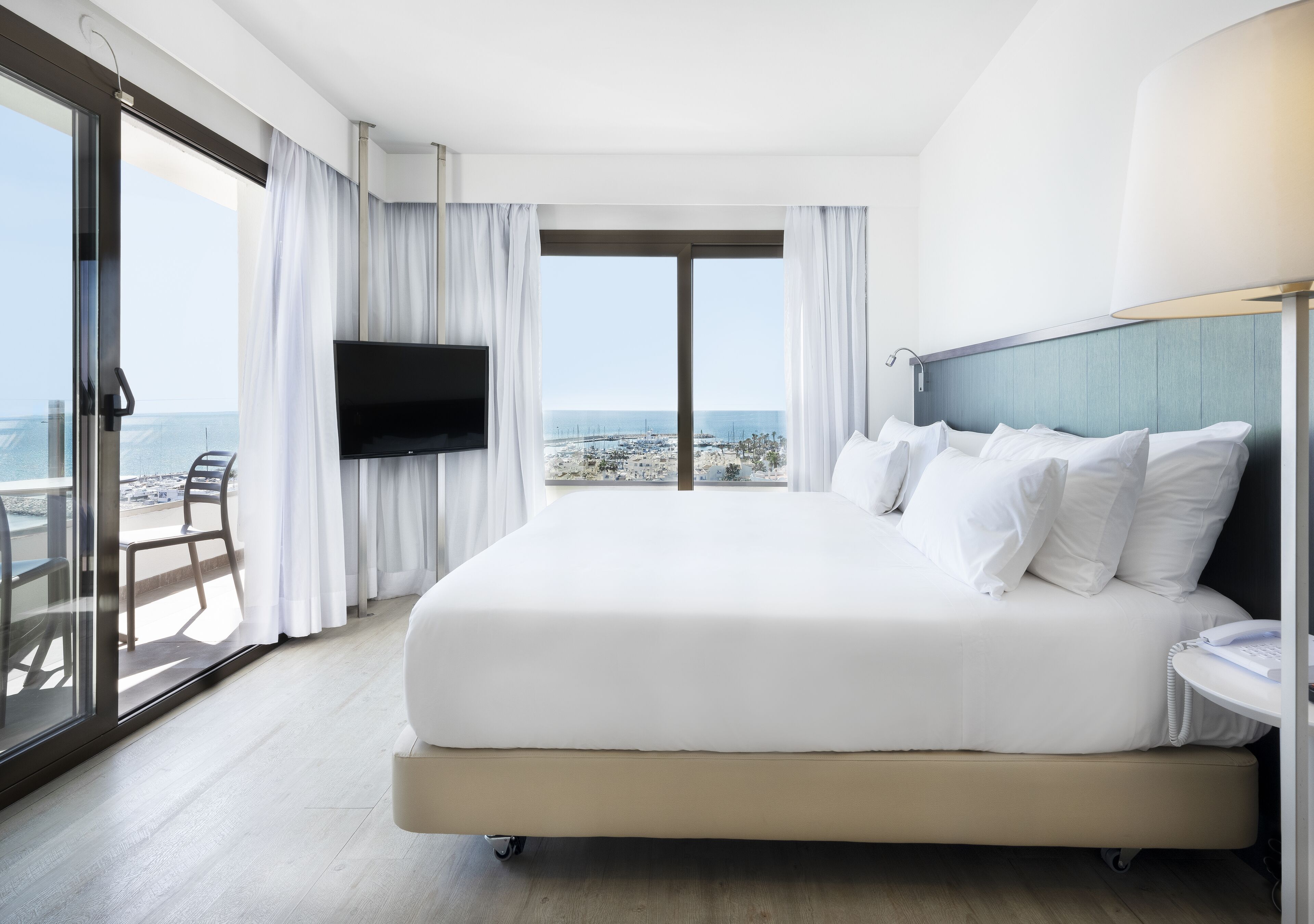 A hotel room with a large bed, a TV, a balcony with a chair, and a view of the city.