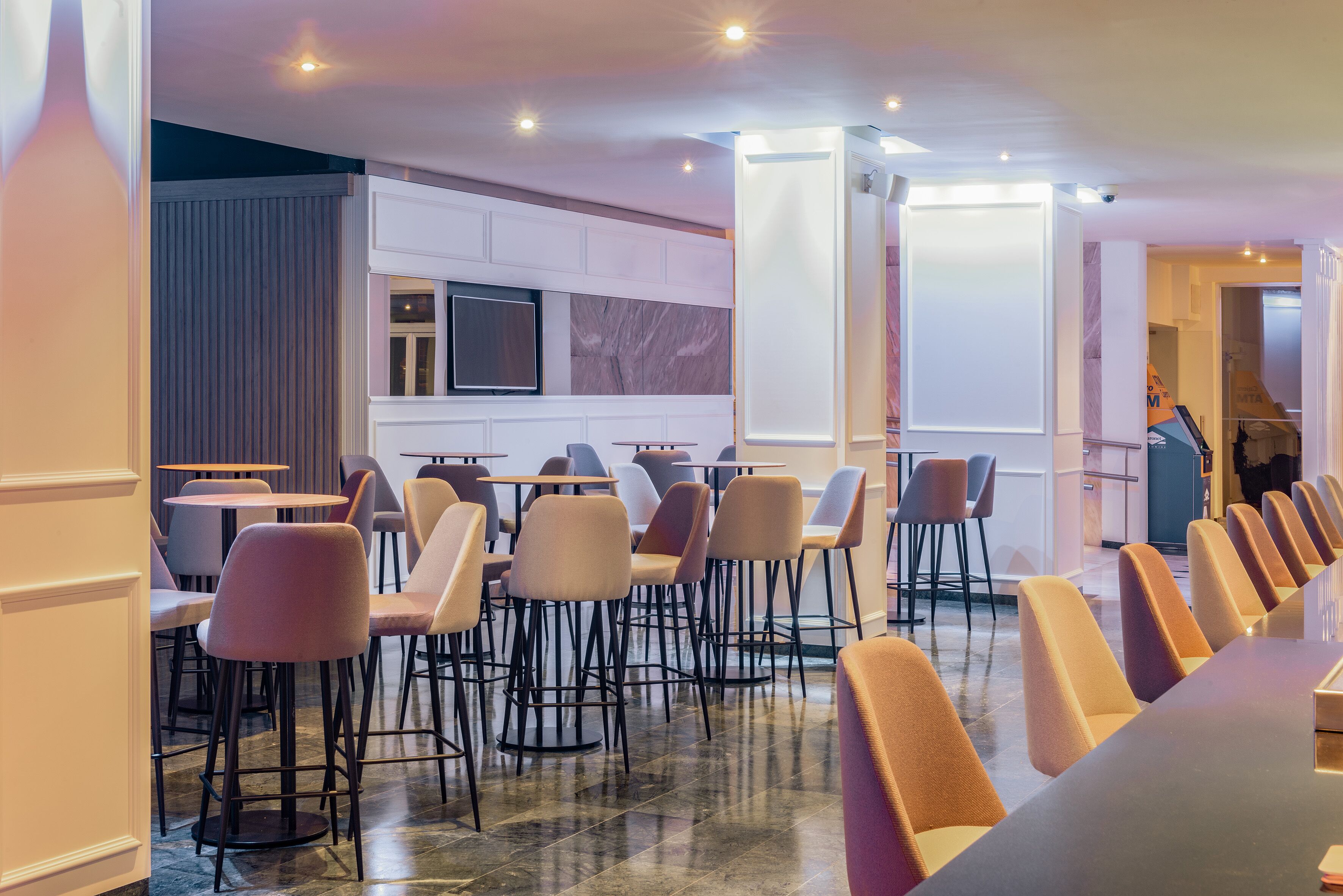 Een moderne hotellobby met een lange rij barstoelen en tafels, een flatscreen televisie aan de muur en een accentmuur van marmer.