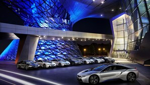 Um edifício moderno com fachada de vidro geométrica singular e uma fileira de carros de luxo estacionados em frente.